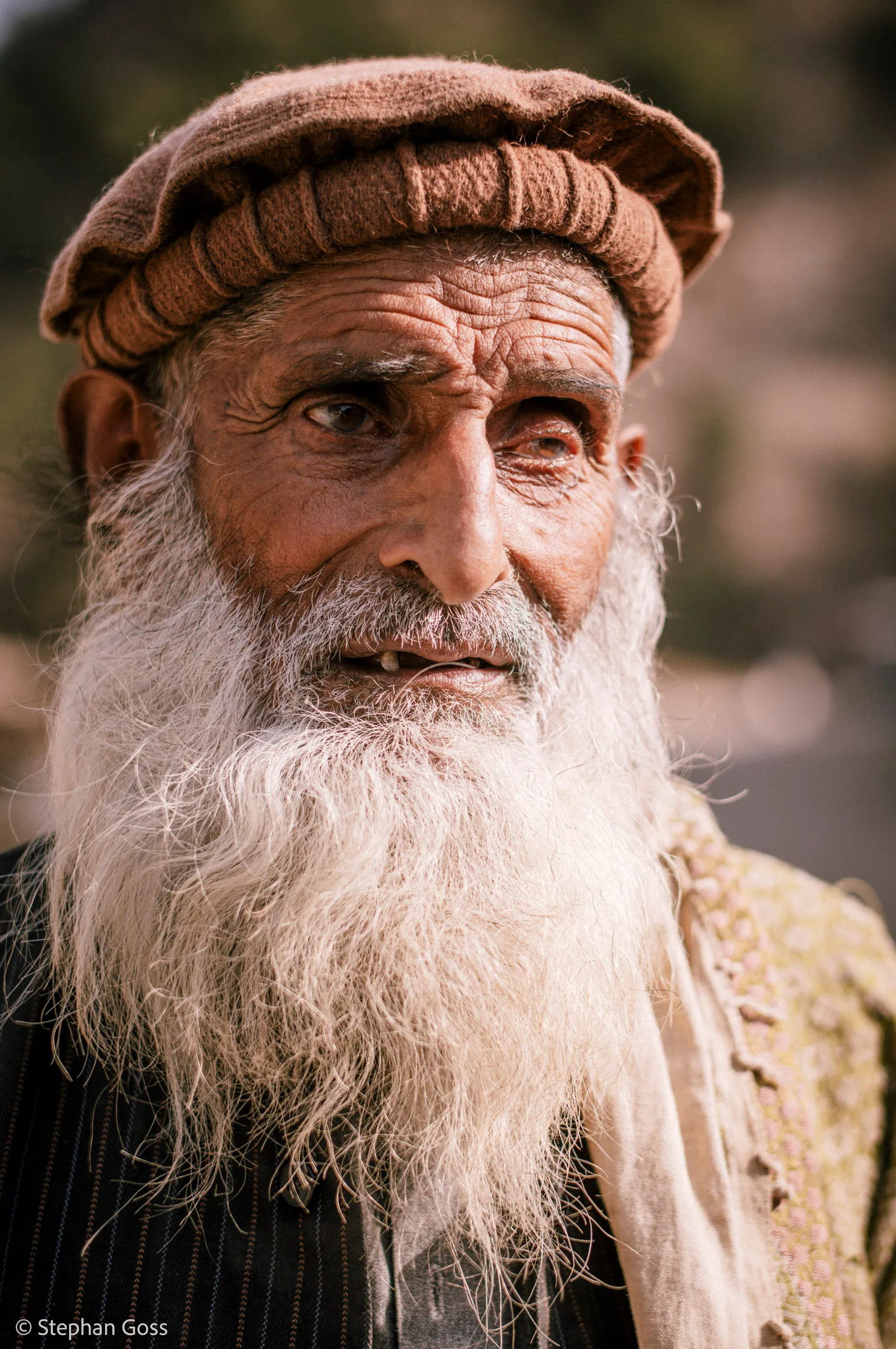 Close portrait of an elderly man with a long white beard and deep-set eyes, wearing a brown wool pakol cap and an embroidered waistcoat over a striped shirt.