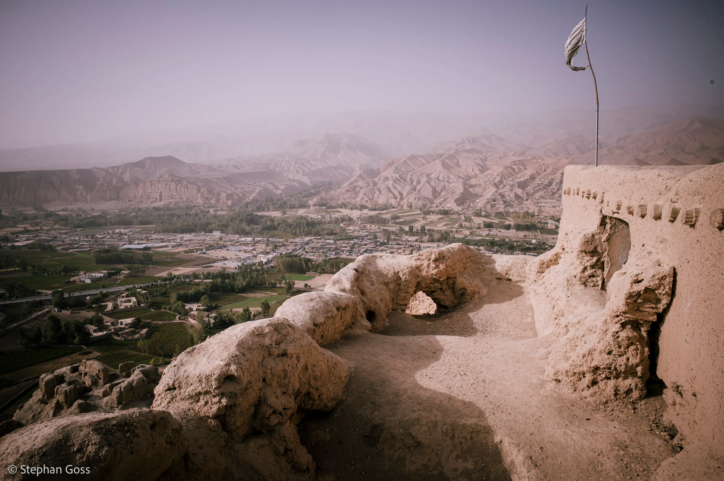 The eroded mud-brick wall of an old hilltop fort in the foreground, a white Taliban flag on a pole at its corner, a green irrigated plain and hazy mountains far below.