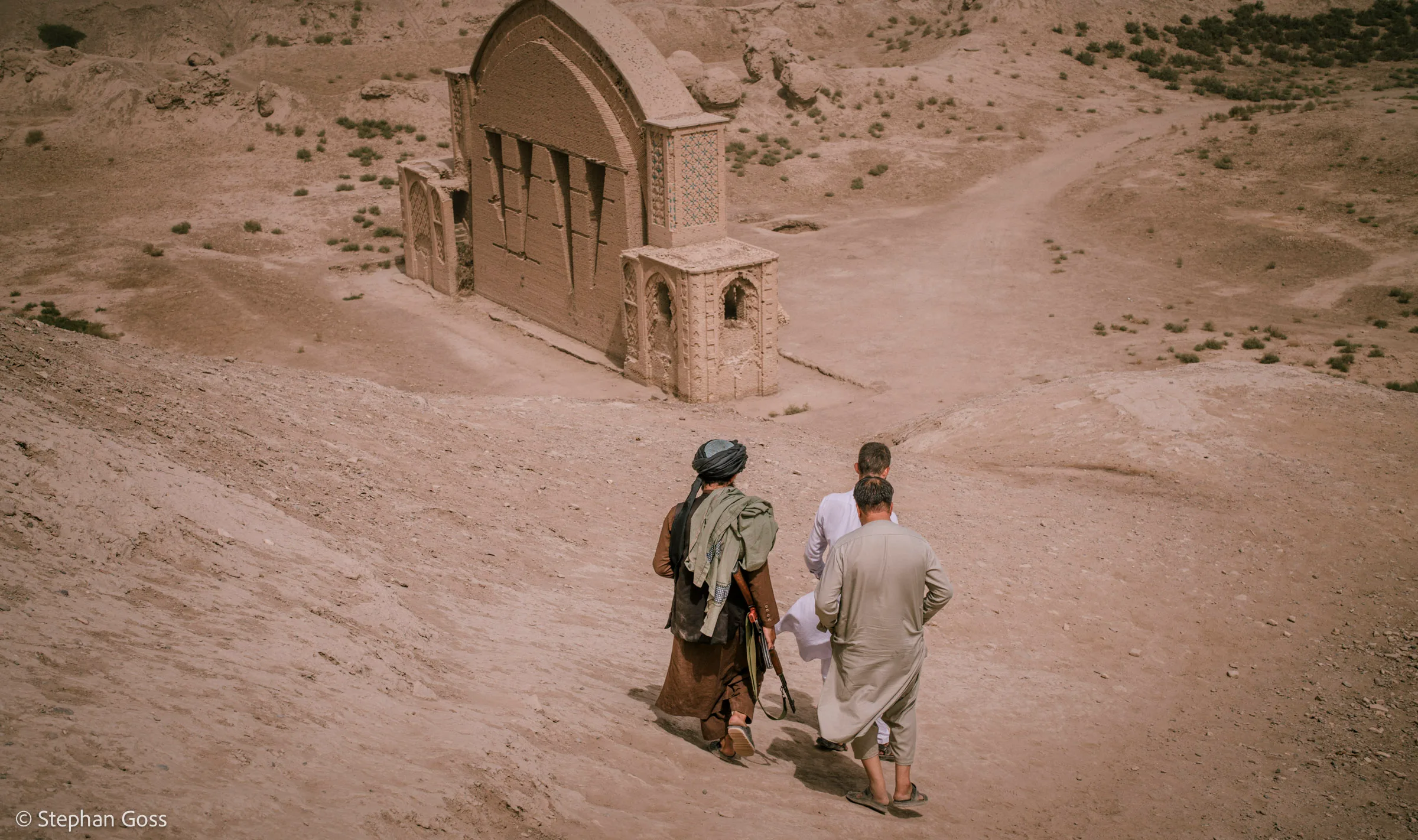 Two men walk down a sandy slope towards a domed brick shrine standing alone among bare desert hills.