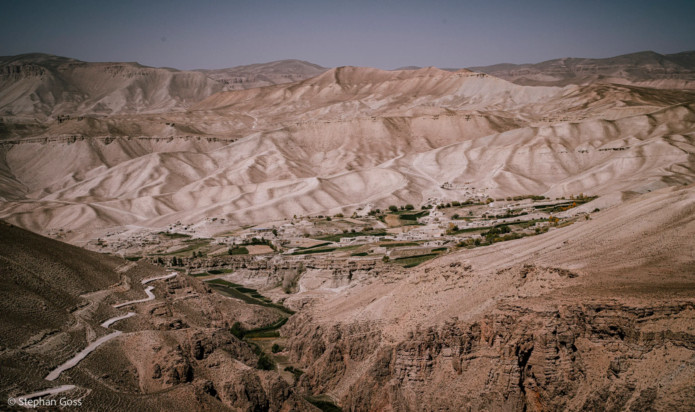 A green ribbon of cultivated land and a scatter of flat-roofed houses follow a river along the floor of a deep, bare valley between eroded desert mountains.