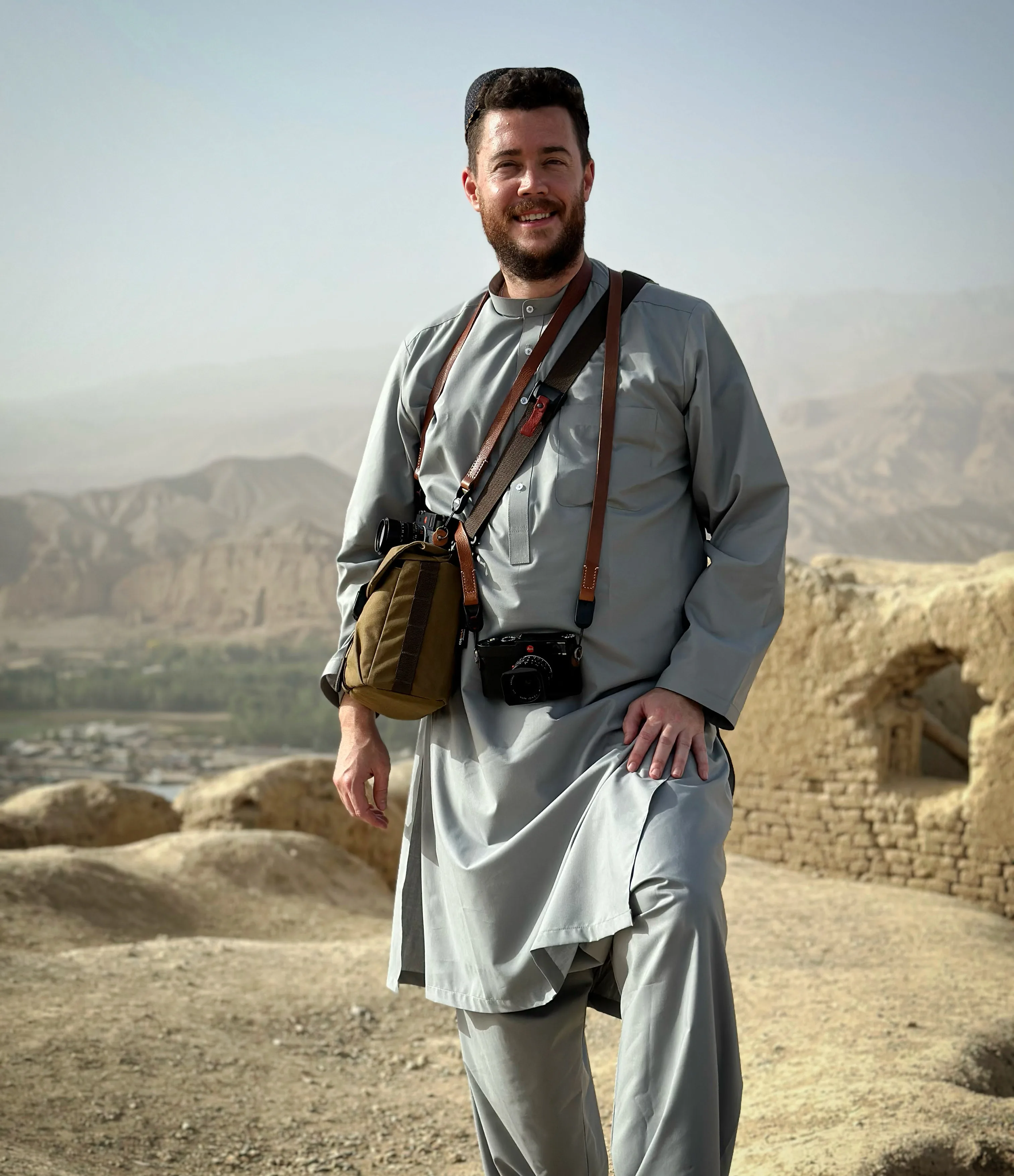 Stephan Goss standing full length on a ridge above a wide valley, in a grey shalwar kameez with a shoulder bag and a camera at his hip.