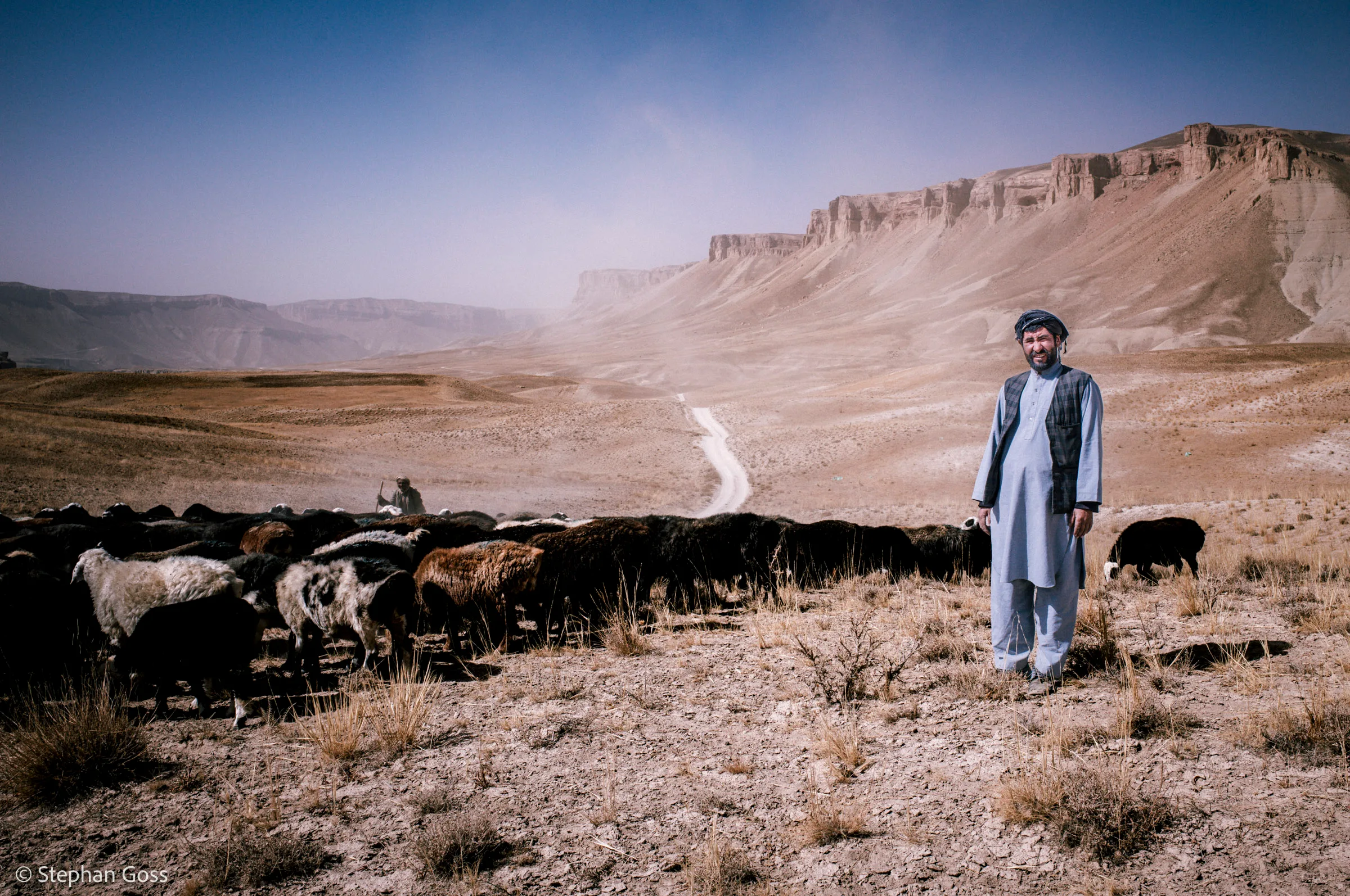 A shepherd in a blue shalwar kameez and turban stands beside his flock of black and brown sheep on a wide dry plain below a long escarpment of pale cliffs.