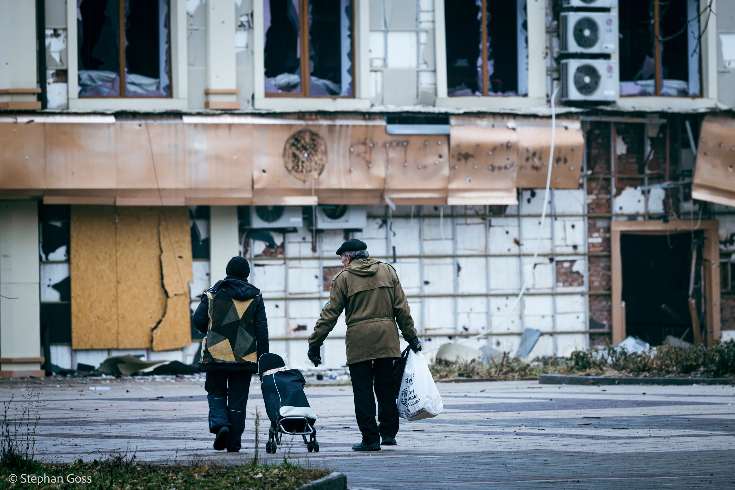 An elderly man carrying a white sack and a woman pulling a shopping trolley walk away from the camera past a shell-damaged building with every window blown out.