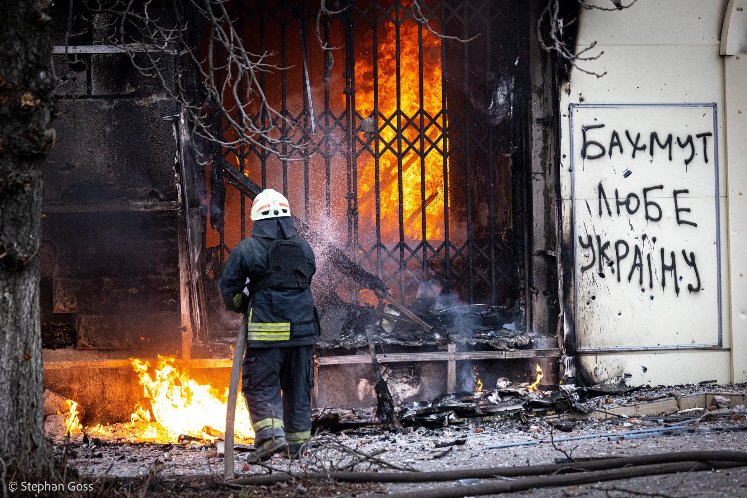 A firefighter directs a hose into a burning shopfront behind iron railings; on the scorched wall beside him someone has painted БАХМУТ ЛЮБЕ УКРАЇНУ in white.