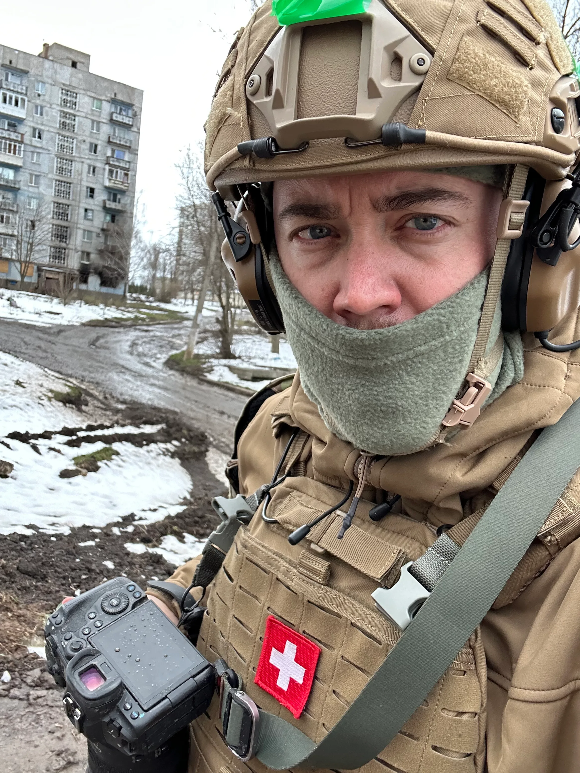 Close portrait of Stephan Goss in a helmet with a neck gaiter pulled up, wearing a plate carrier with a Swiss flag patch and a camera on a sling, on a snowy street lined with apartment blocks.