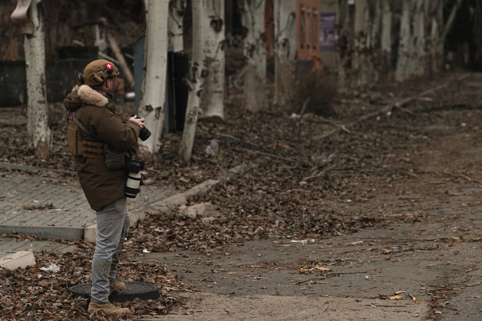 A lone figure in a brown parka stands photographing across a bare, debris-strewn street lined with leafless trees.
