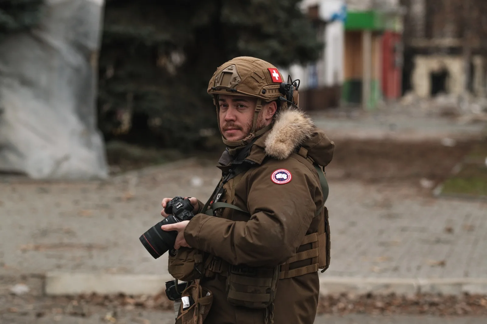 Stephan Goss in a helmet with a Swiss flag patch and a fur-trimmed parka, camera held at his chest, a damaged street behind him.