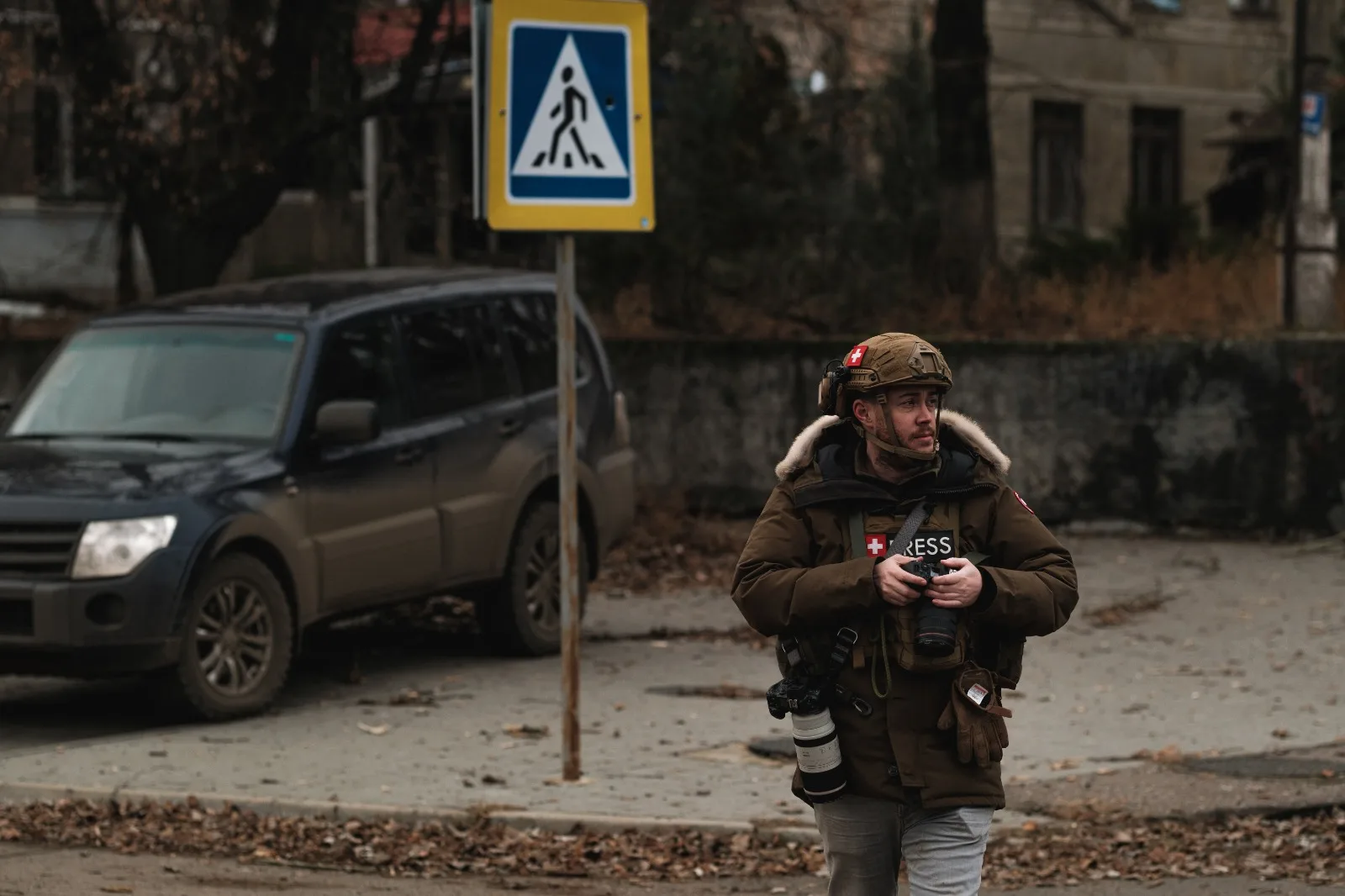 Stephan Goss walking along a road in press body armour, past a parked vehicle and a pedestrian-crossing sign, with damaged buildings behind.