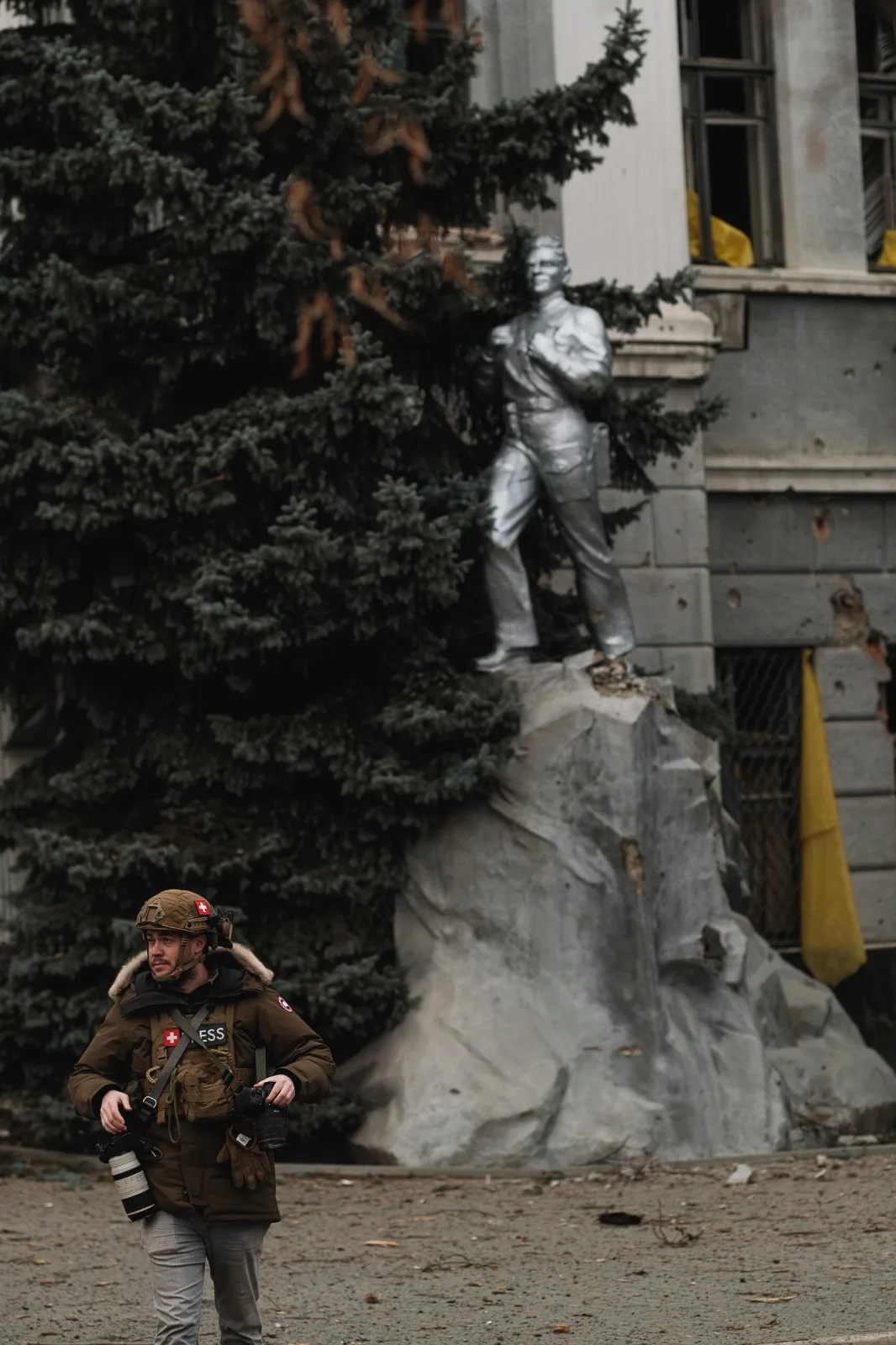A small figure in body armour walks past a large stone monument standing in front of a damaged building.