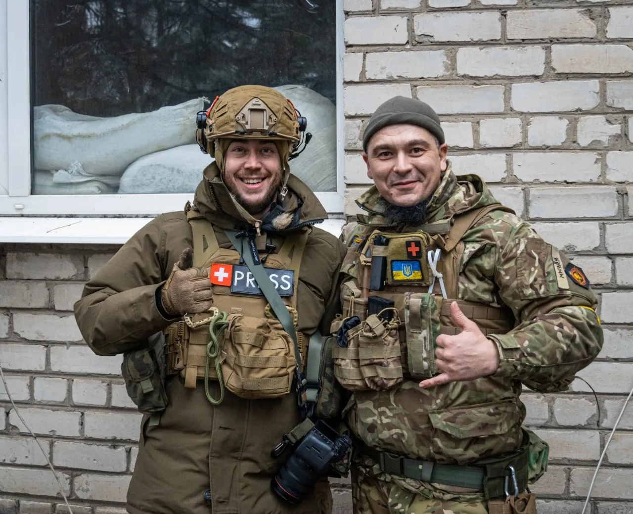 Stephan Goss and a Ukrainian soldier standing side by side against a white brick wall, both smiling. He wears a helmet and a plate carrier marked PRESS; the soldier is in camouflage.