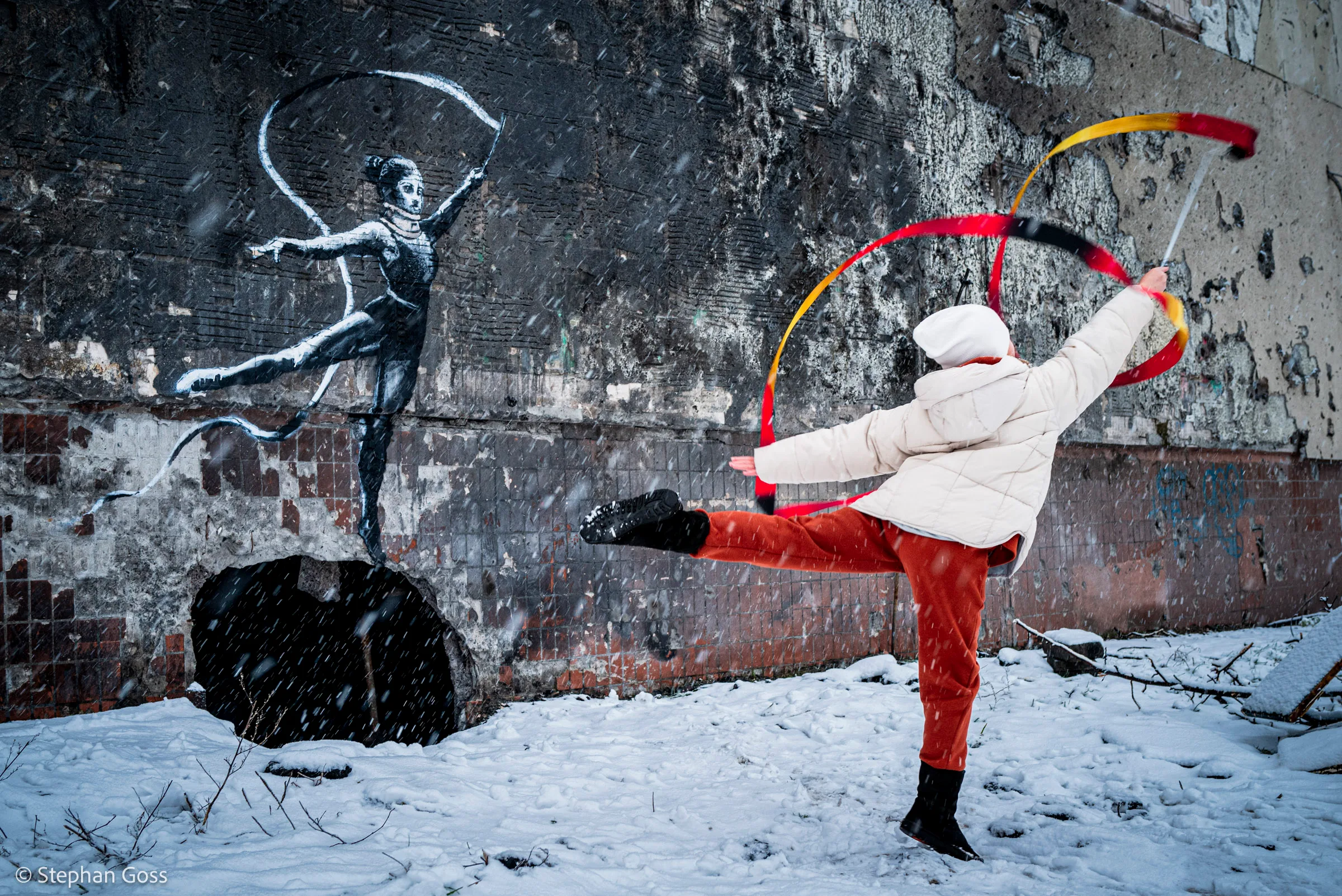 A girl in a white padded coat and red trousers dances with a ribbon in falling snow in front of a Banksy stencil of a gymnast painted on a fire-blackened wall.