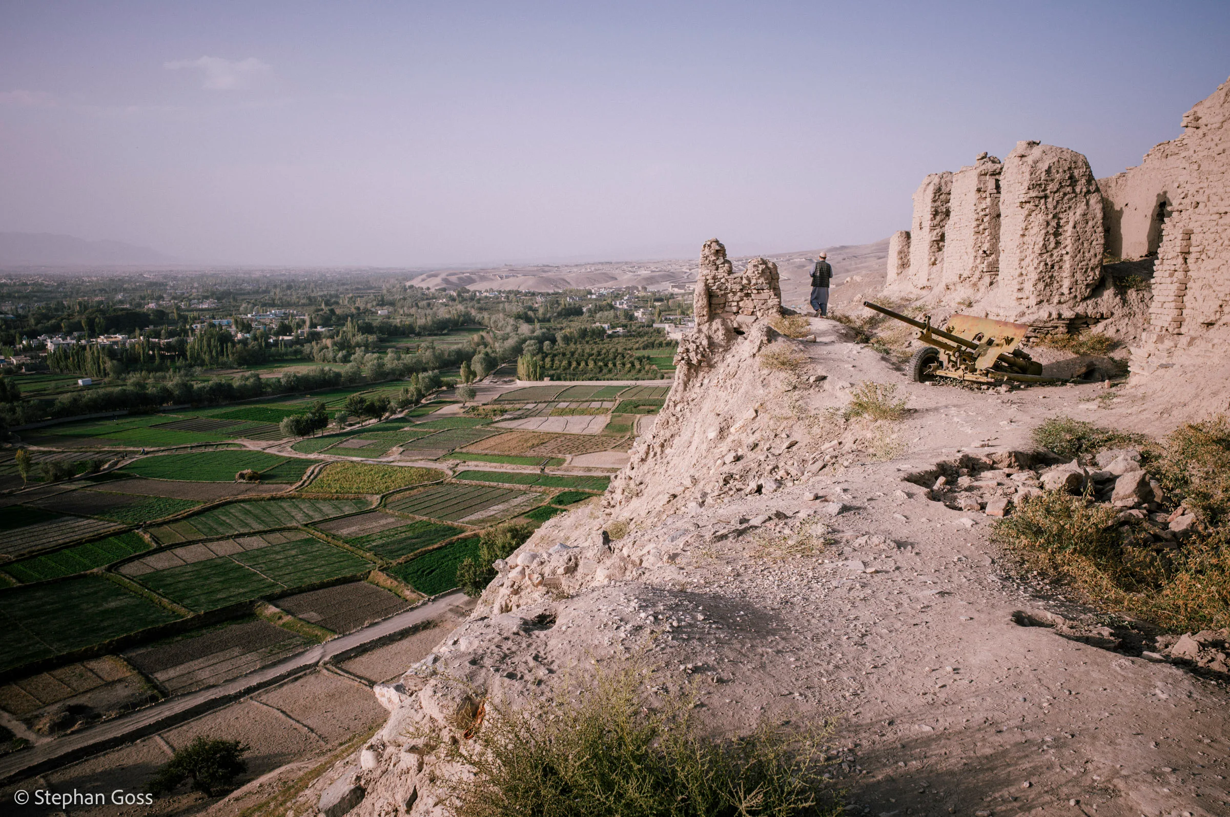 A Soviet-era artillery piece sits among crumbling mud fortifications on a ridge, two figures standing beside it, a patchwork of green fields on the plain far below.