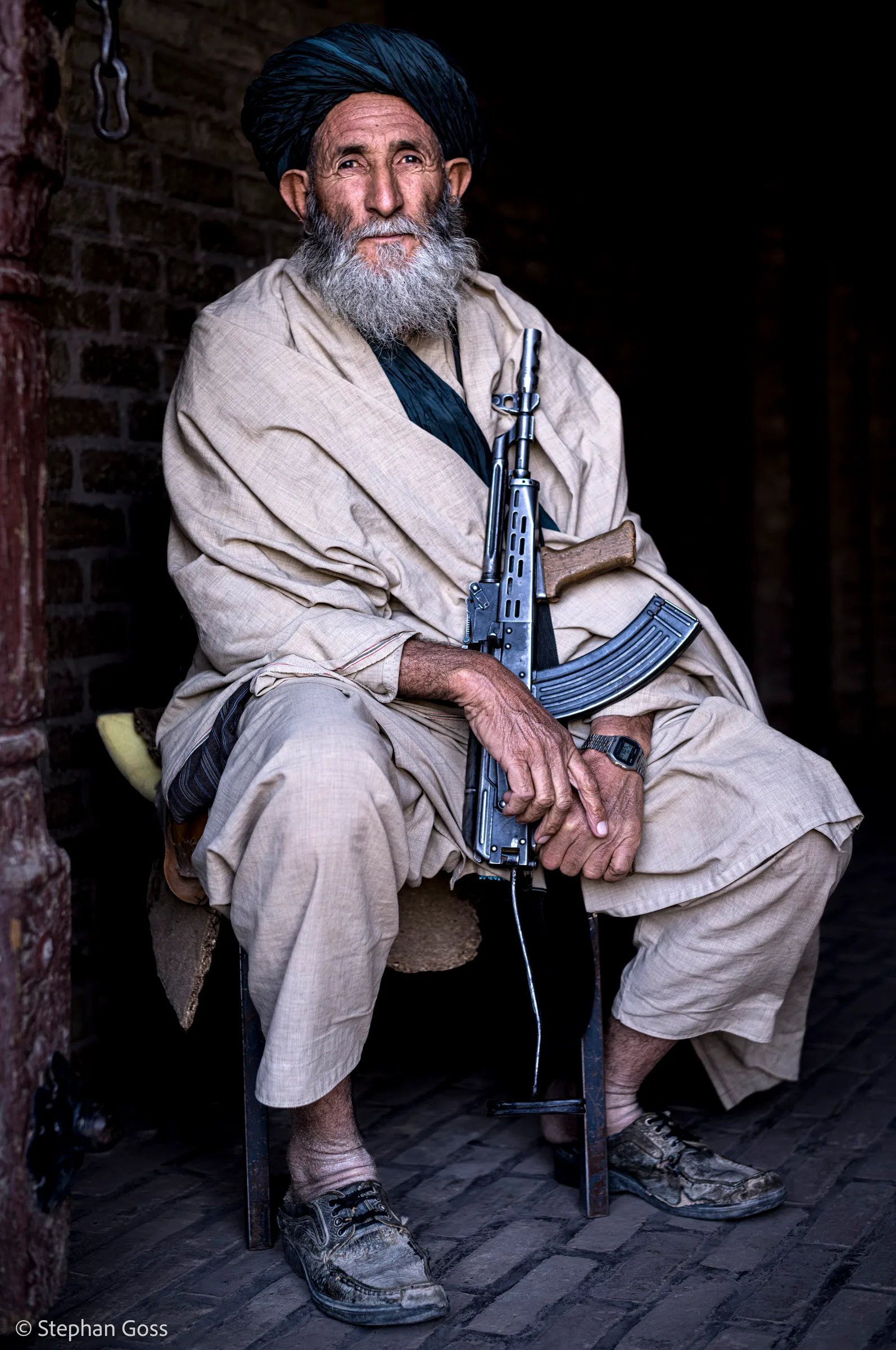 An elderly man with a white beard and dark turban sits on a low stool in a brick doorway, a rifle upright between his knees and both hands resting on it.