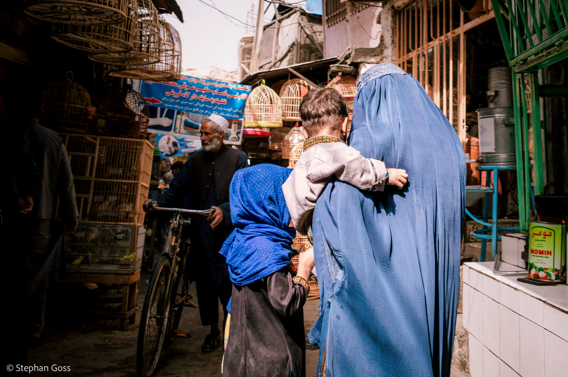 A woman in a blue burqa carries a small child on her hip through a lane hung with wooden and wire birdcages, a girl in a blue headscarf at her side and a man with a bicycle behind.