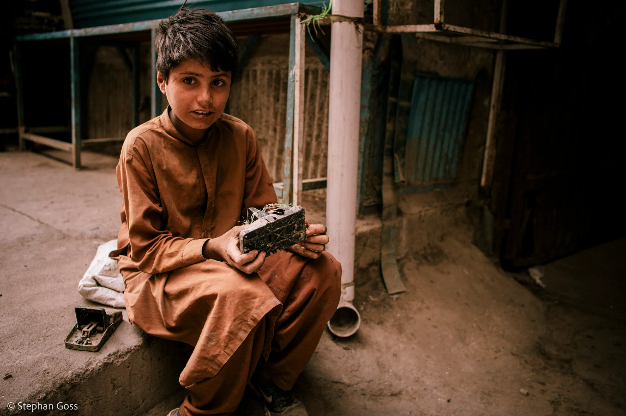 A boy of about ten in a rust-coloured shalwar kameez sits on a kerb holding a heavy iron padlock, a second lock and a cloth bundle on the step beside him.