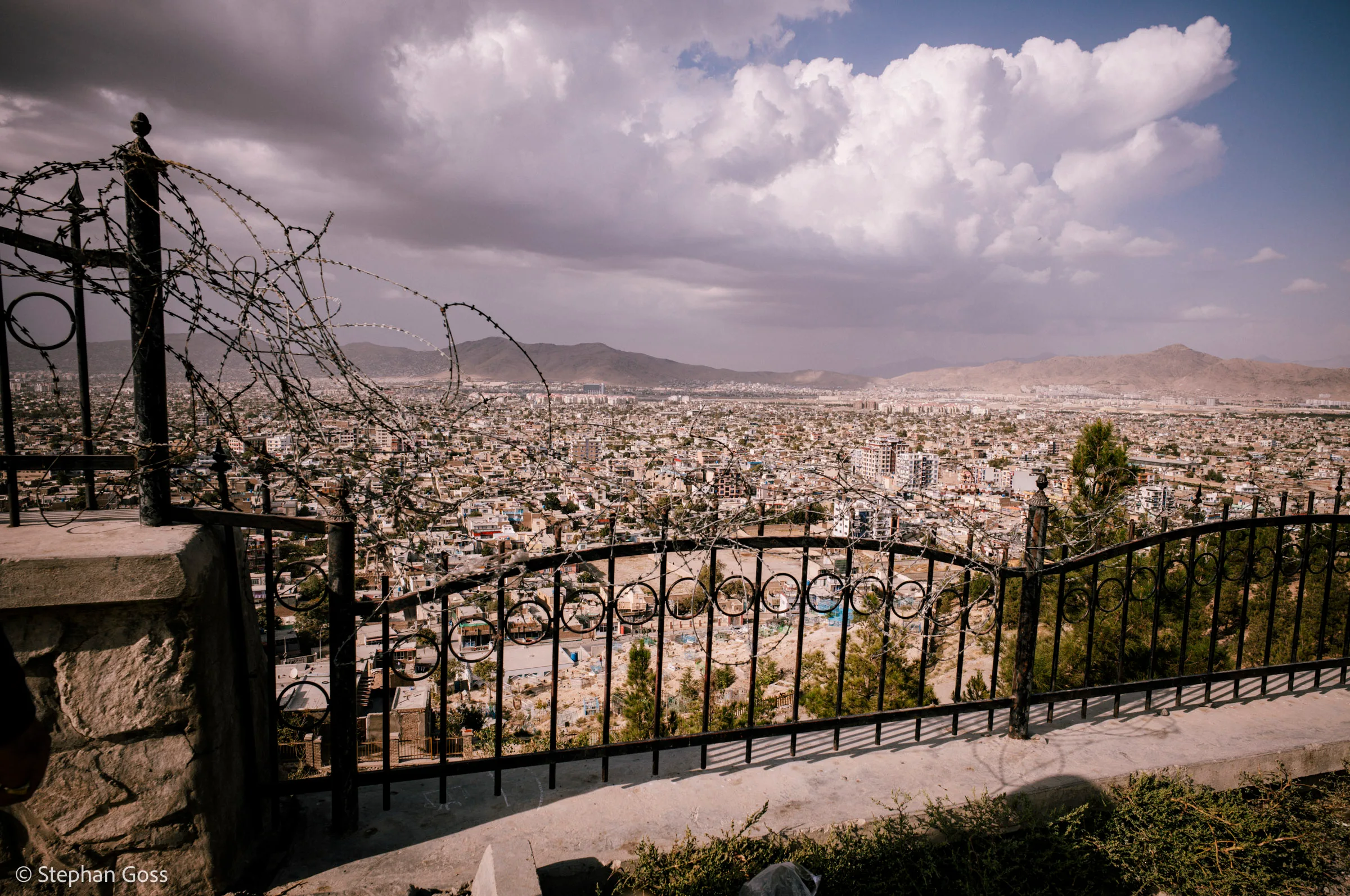A city spread across its plain under towering white cloud, seen from a hillside through an ornamental iron railing topped with coils of razor wire.