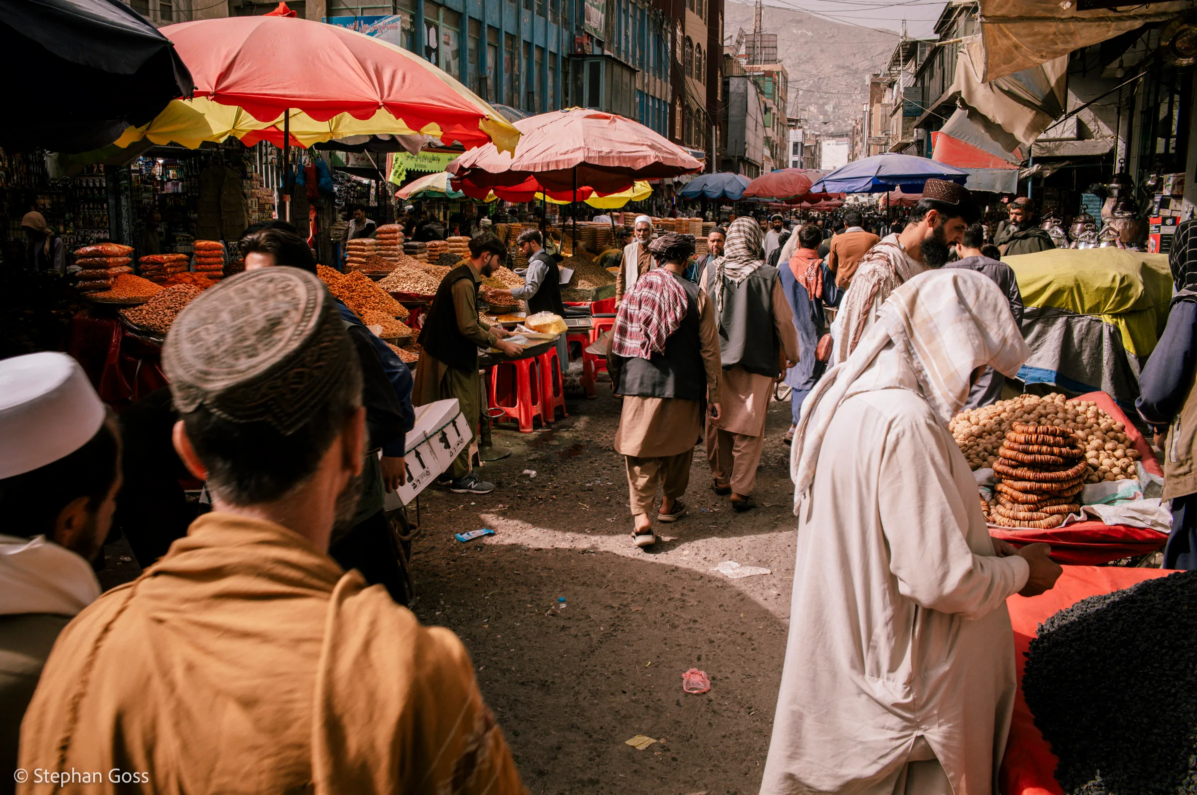 A crowded bazaar street at eye level: men in shalwar kameez move between stalls piled with bread rings and dried fruit under red and yellow parasols, hills rising behind.
