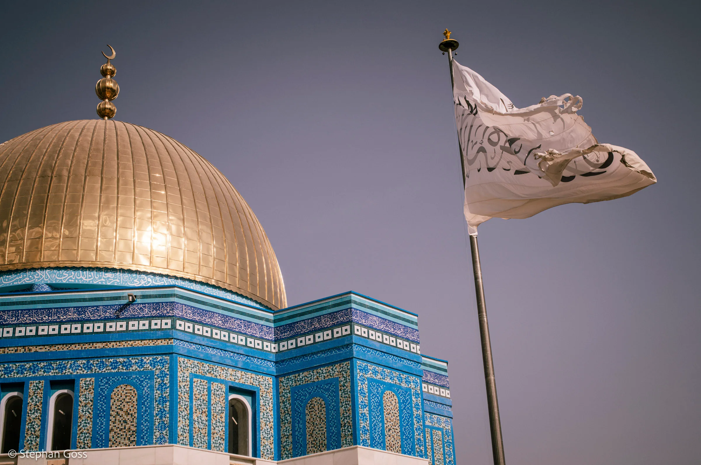 The gilded dome and blue-tiled drum of a shrine against a dusk sky, with a large white Taliban flag flying from a mast beside it.