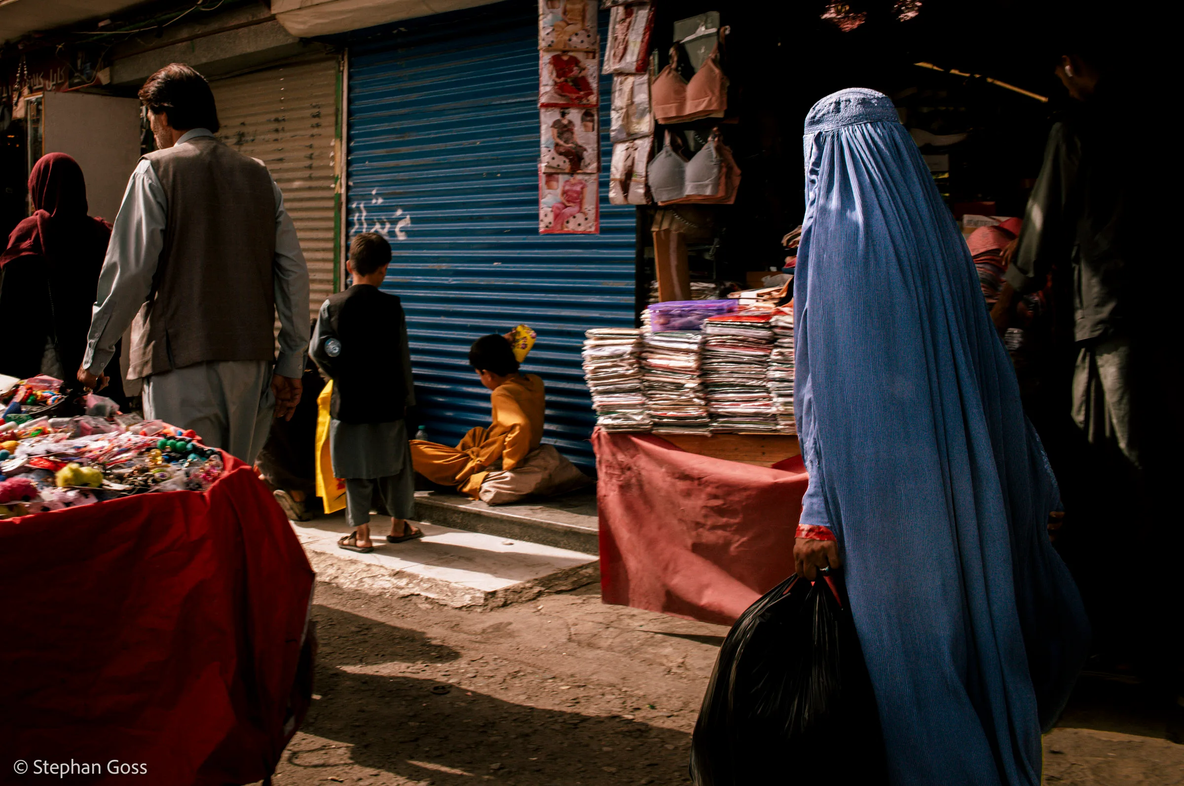 A woman in a blue burqa carrying a black plastic bag walks through a narrow market street past haberdashery stalls; two children sit on the kerb in a patch of sun.