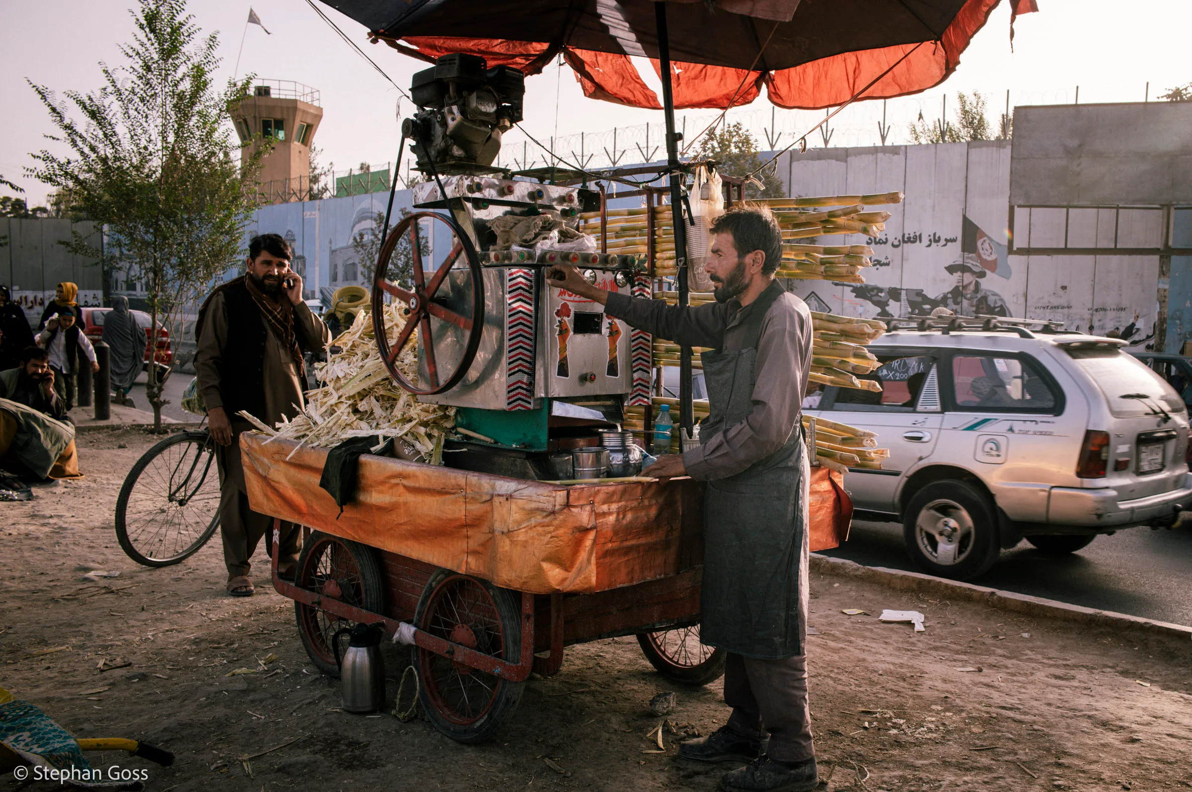 A man works a hand-cranked sugarcane press mounted on a wooden handcart under a red parasol at dusk, bundles of cane stacked behind him and a guard tower on the wall beyond.