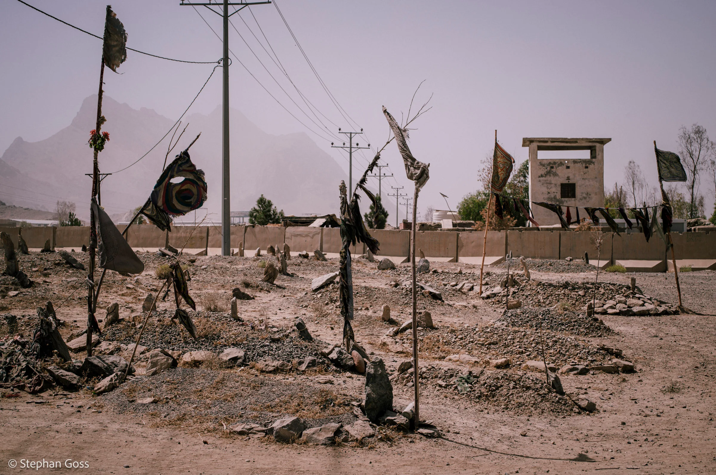 A graveyard of low stone-covered mounds on bare ground, each marked with a tattered flag on a thin pole, a monument and mountains behind in the dust haze.