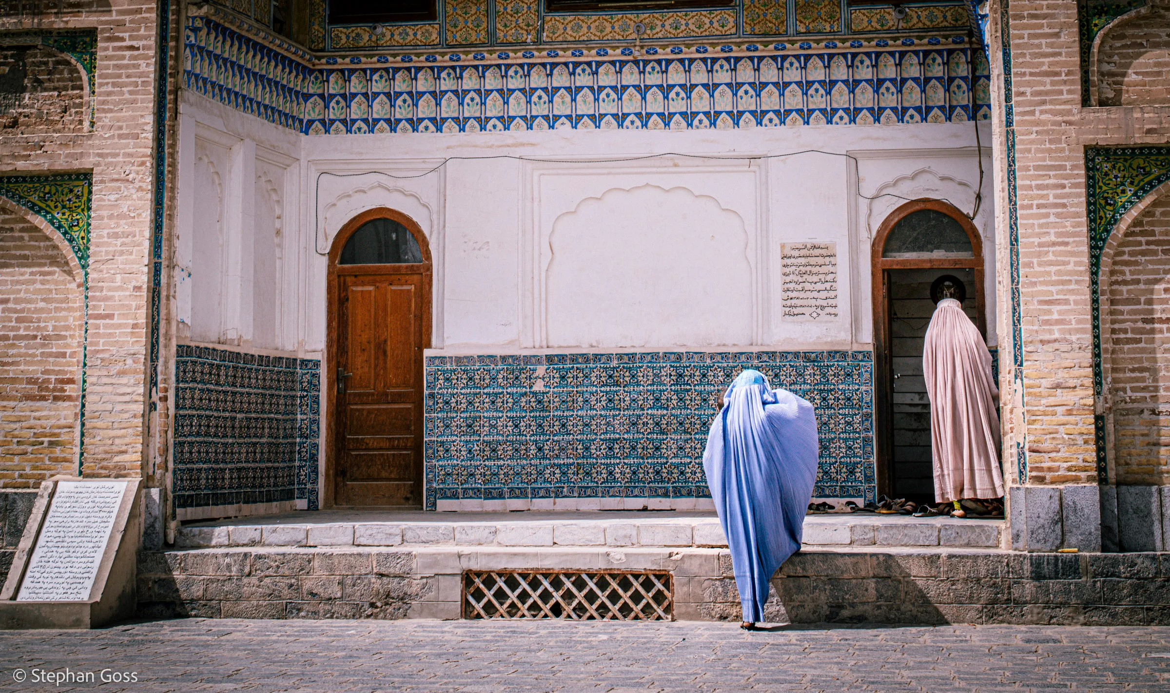 Two women, one in a pale blue burqa and one in a cream chador, cross a tiled courtyard towards separate doorways of a mosque decorated in blue and gold.