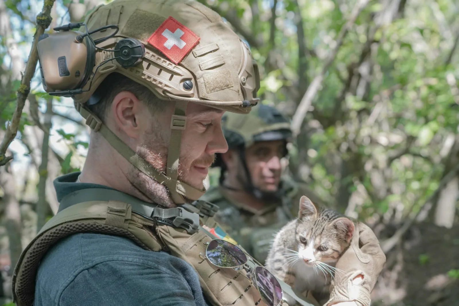 Stephan Goss in a helmet and body armour holding a small tabby cat in the crook of his arm, a soldier standing among trees behind him.