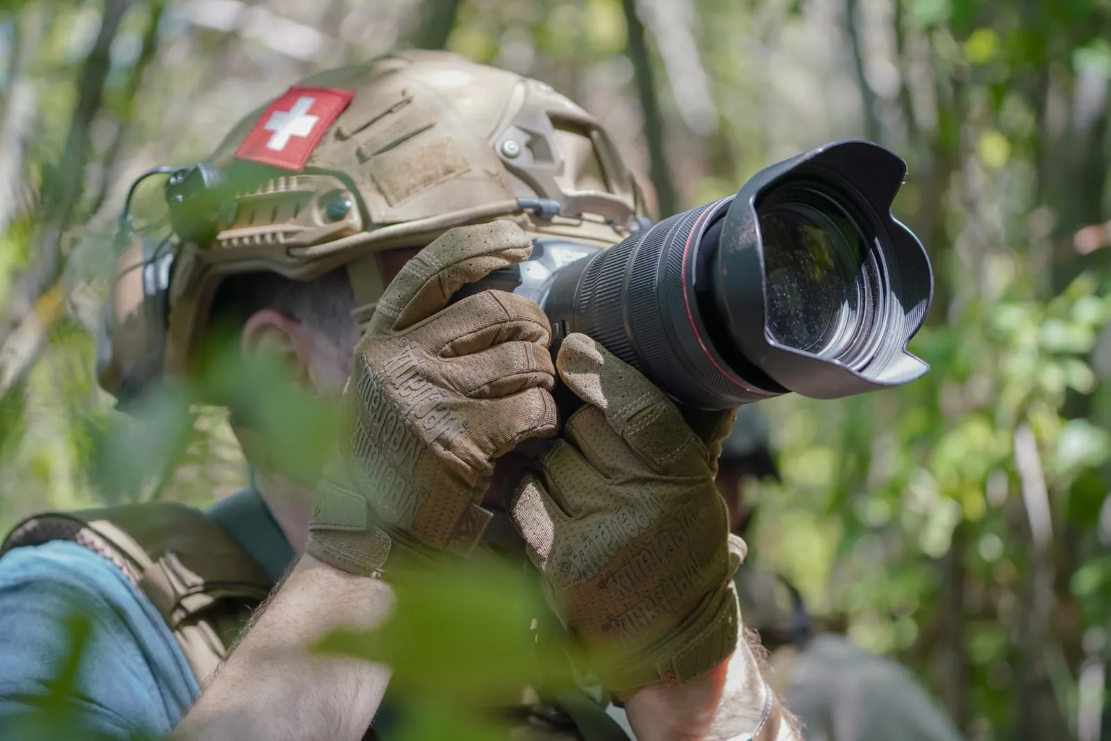 Gloved hands raising a camera with a long telephoto lens to the eye, the photographer in a helmet with a Swiss flag patch, foliage across the foreground.