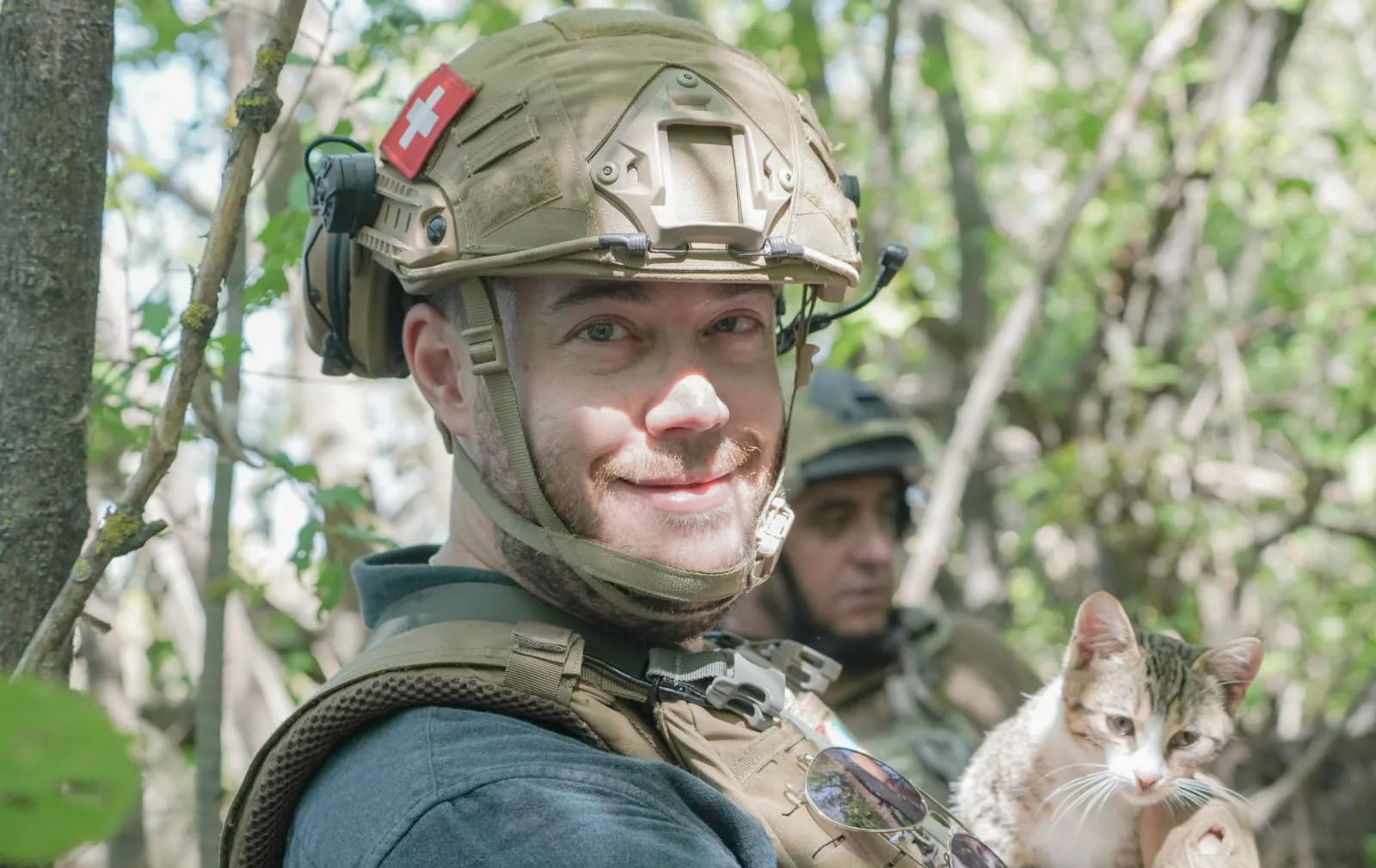 Stephan Goss looking at the camera and smiling, in a helmet with a Swiss flag patch, holding a small tabby cat, with a soldier behind him among trees.