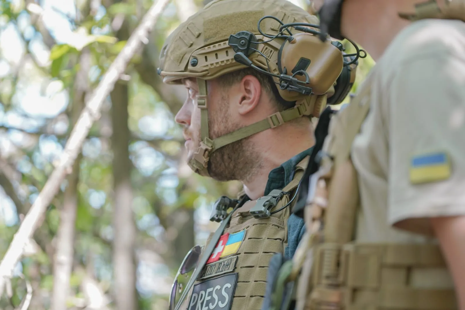 Stephan Goss in profile in a helmet and headset, wearing a plate carrier marked PRESS with a Ukrainian flag patch, facing a soldier whose sleeve also carries a Ukrainian flag.