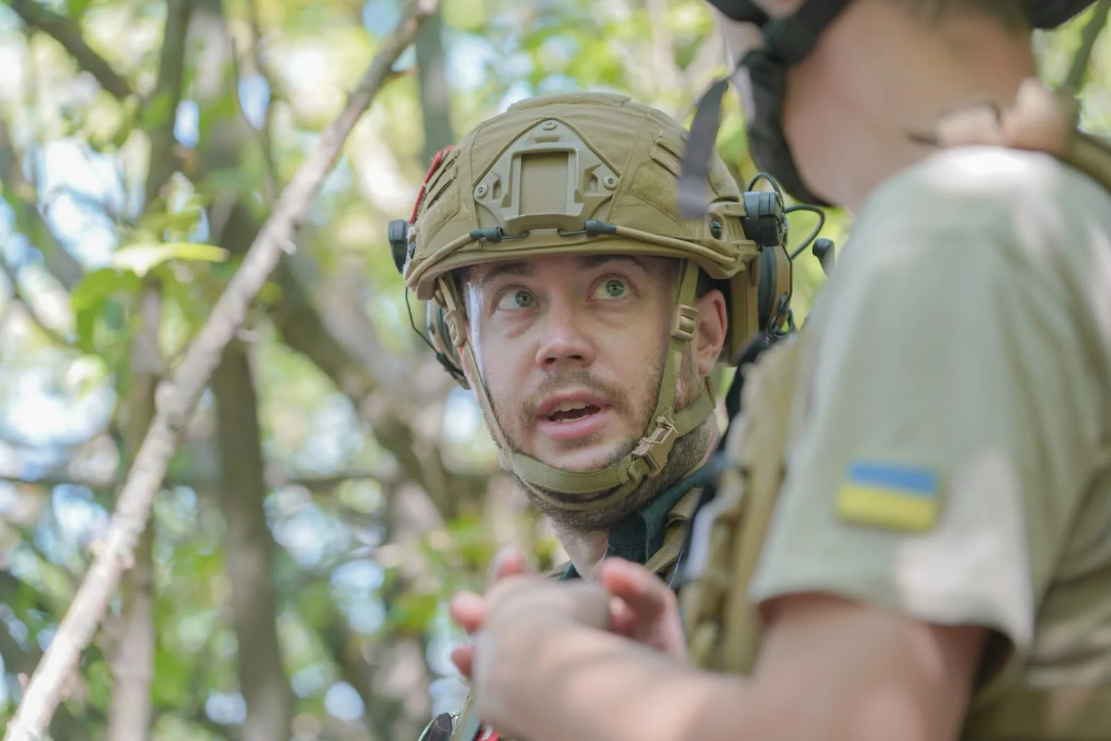 Stephan Goss in a helmet and headset looking towards the camera, with a soldier’s shoulder and Ukrainian flag patch in the foreground.