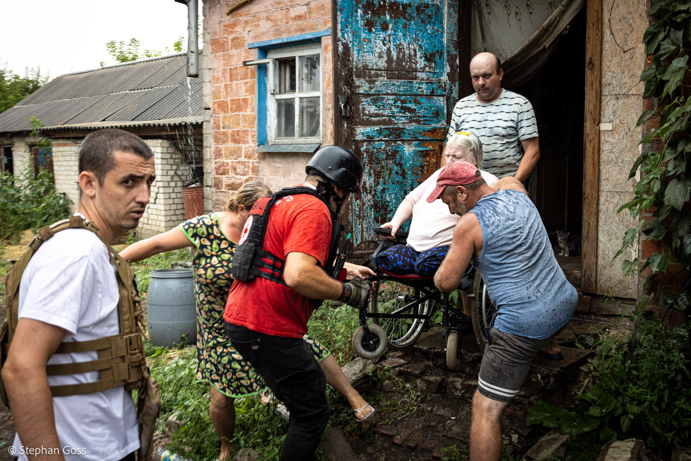 Volunteers and neighbours lift a woman in a wheelchair down the steps of a village house, one in a helmet and red vest, another in body armour watching the road.