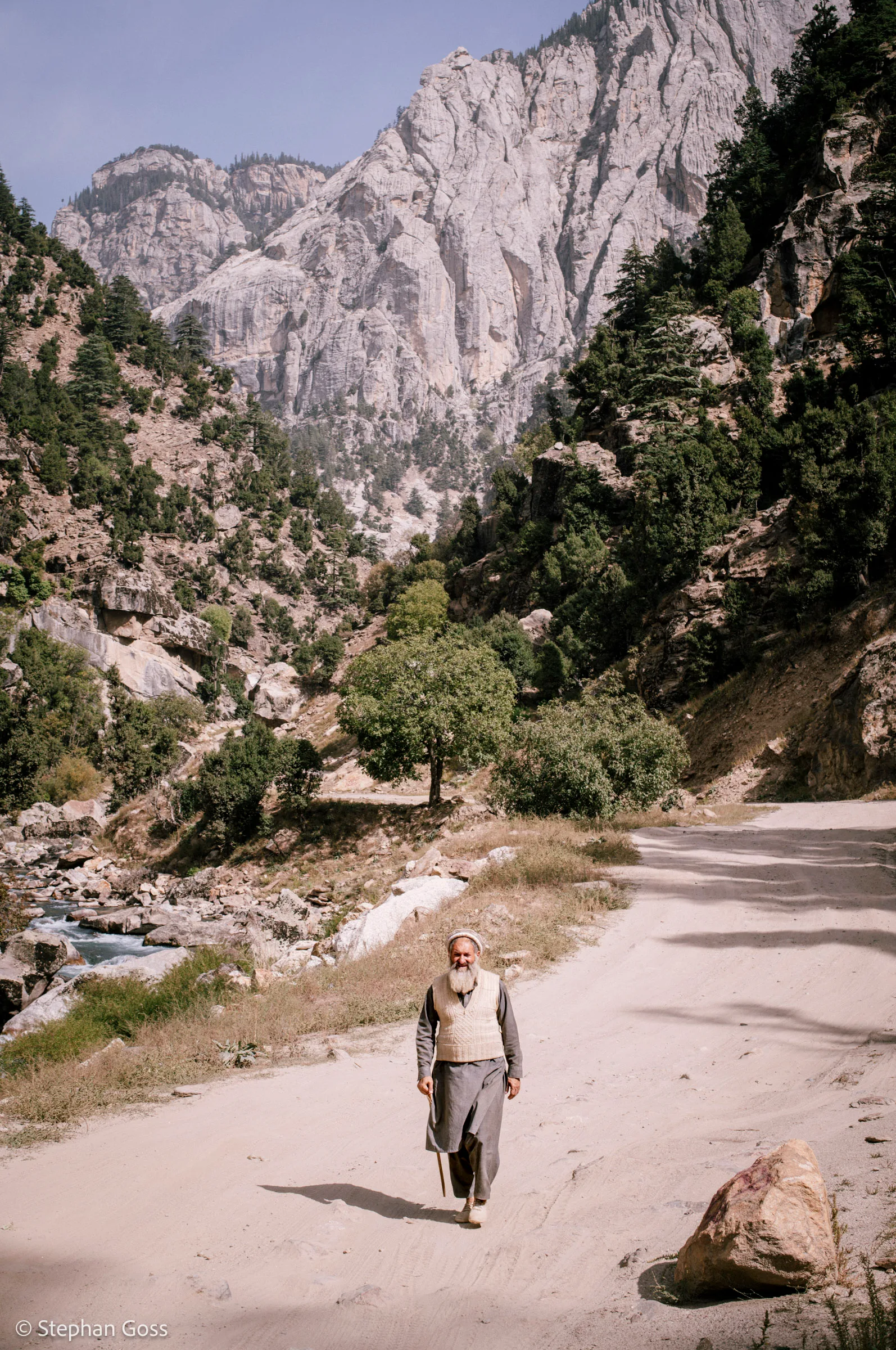 A man in a waistcoat and cap walks up the middle of a pale dirt road towards the camera, granite peaks and pine forest rising steeply on both sides.