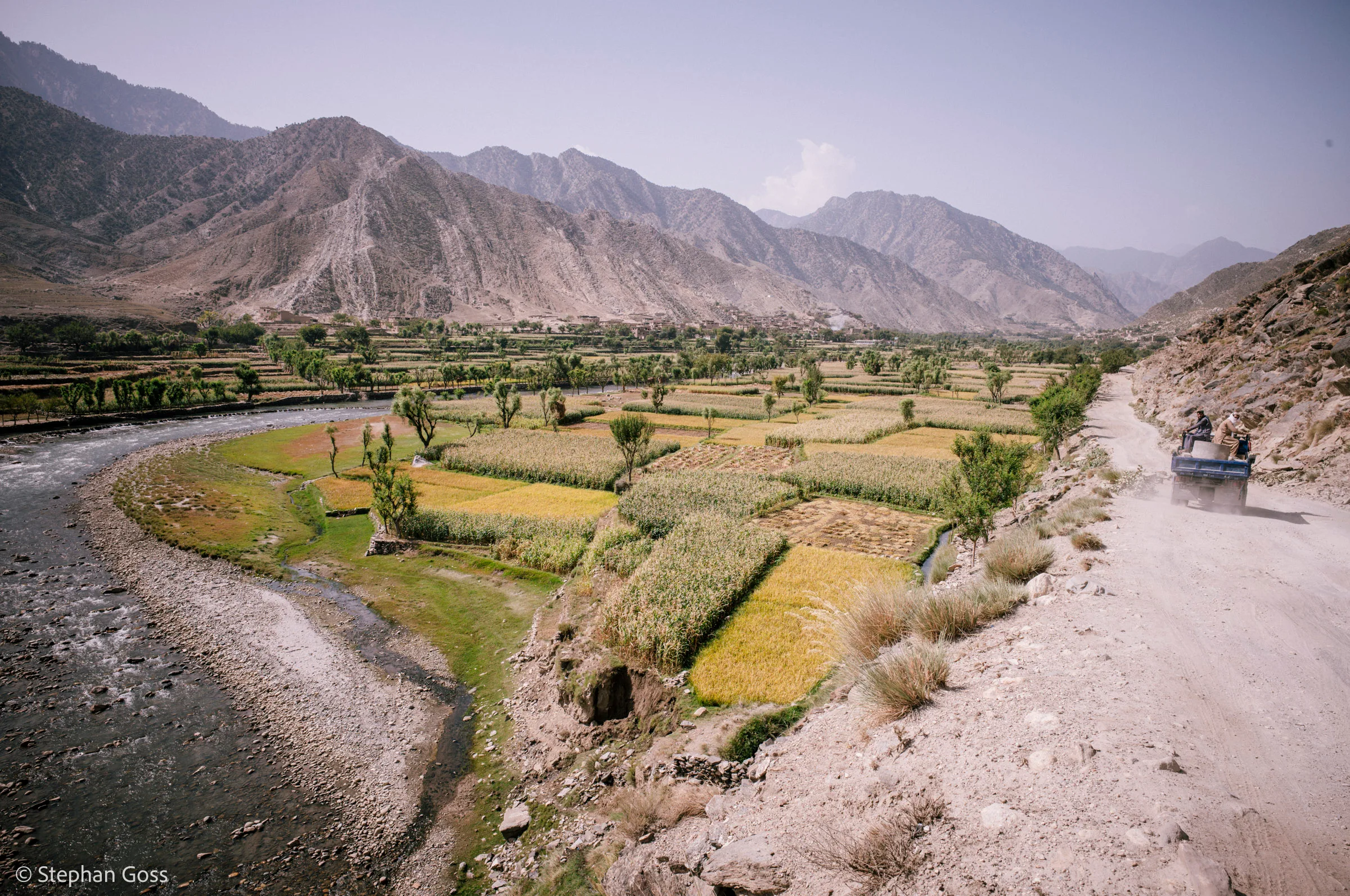 Terraced fields of ripening maize and rice follow a river through a steep mountain valley, a dirt road cut into the slope on the right with a vehicle on it.