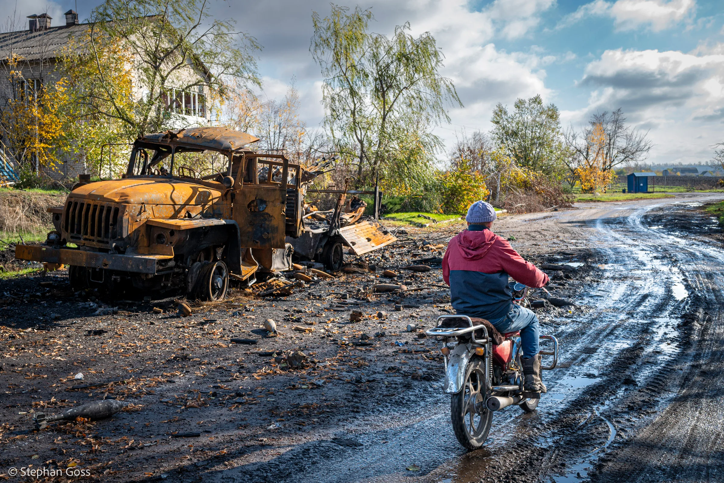 A man on a motorcycle rides along a wet, muddy road past the burnt-out wreck of a military truck outside a village house.
