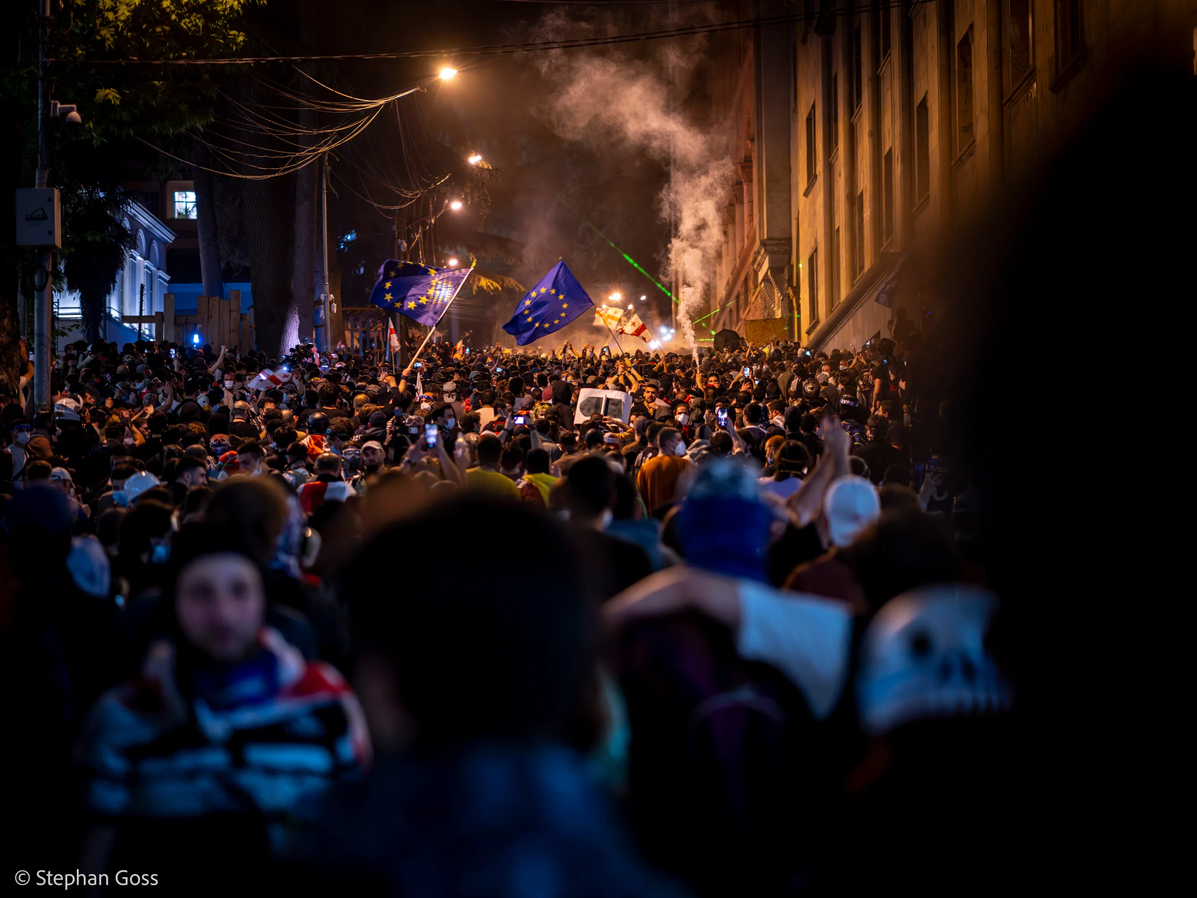 A vast crowd fills a boulevard at night, European Union flags raised above it and smoke drifting through the light of the street lamps.