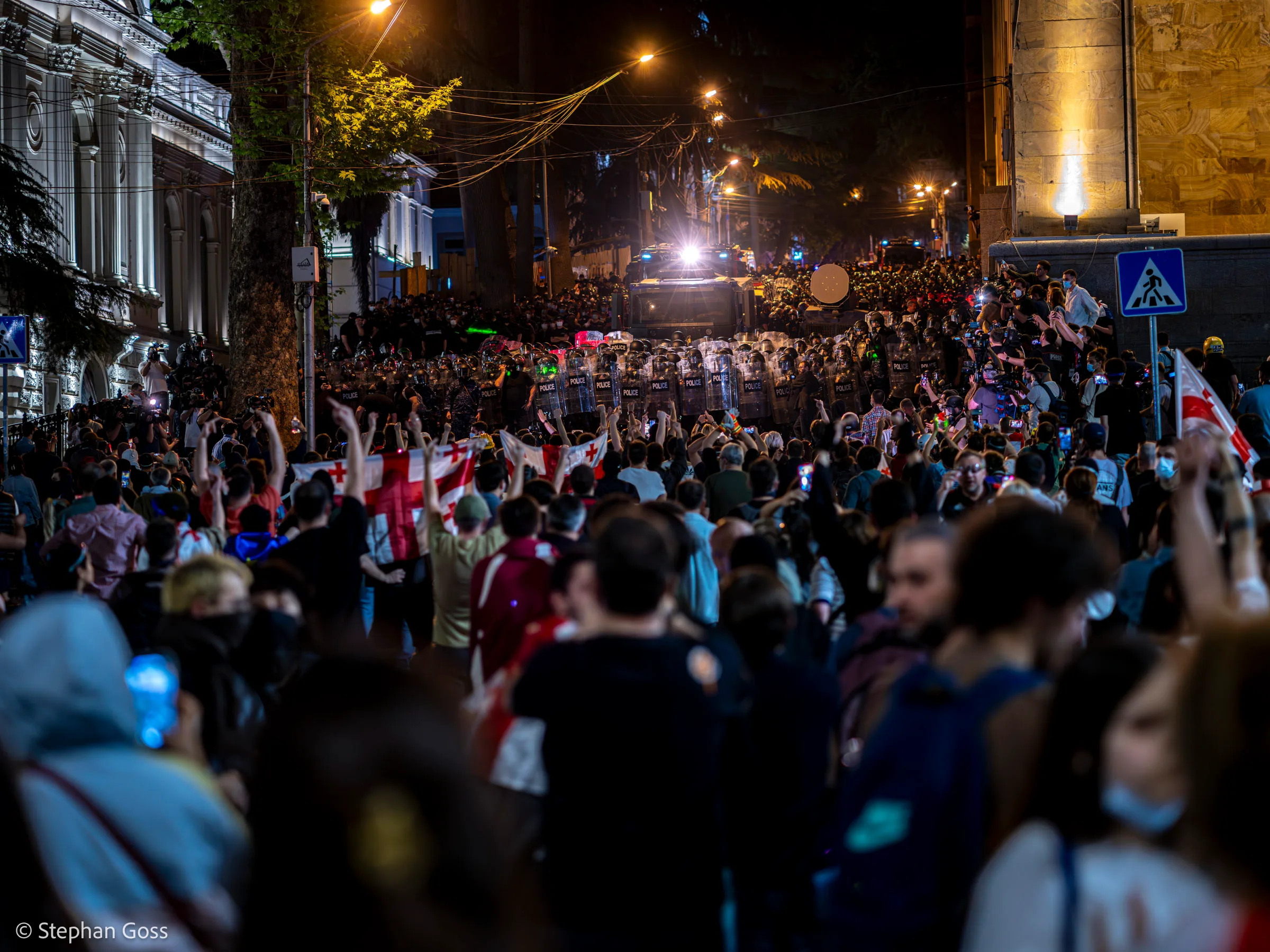 A dense crowd carrying Georgian flags fills a street at night, facing a line of riot police with shields and a water cannon truck drawn up behind them.