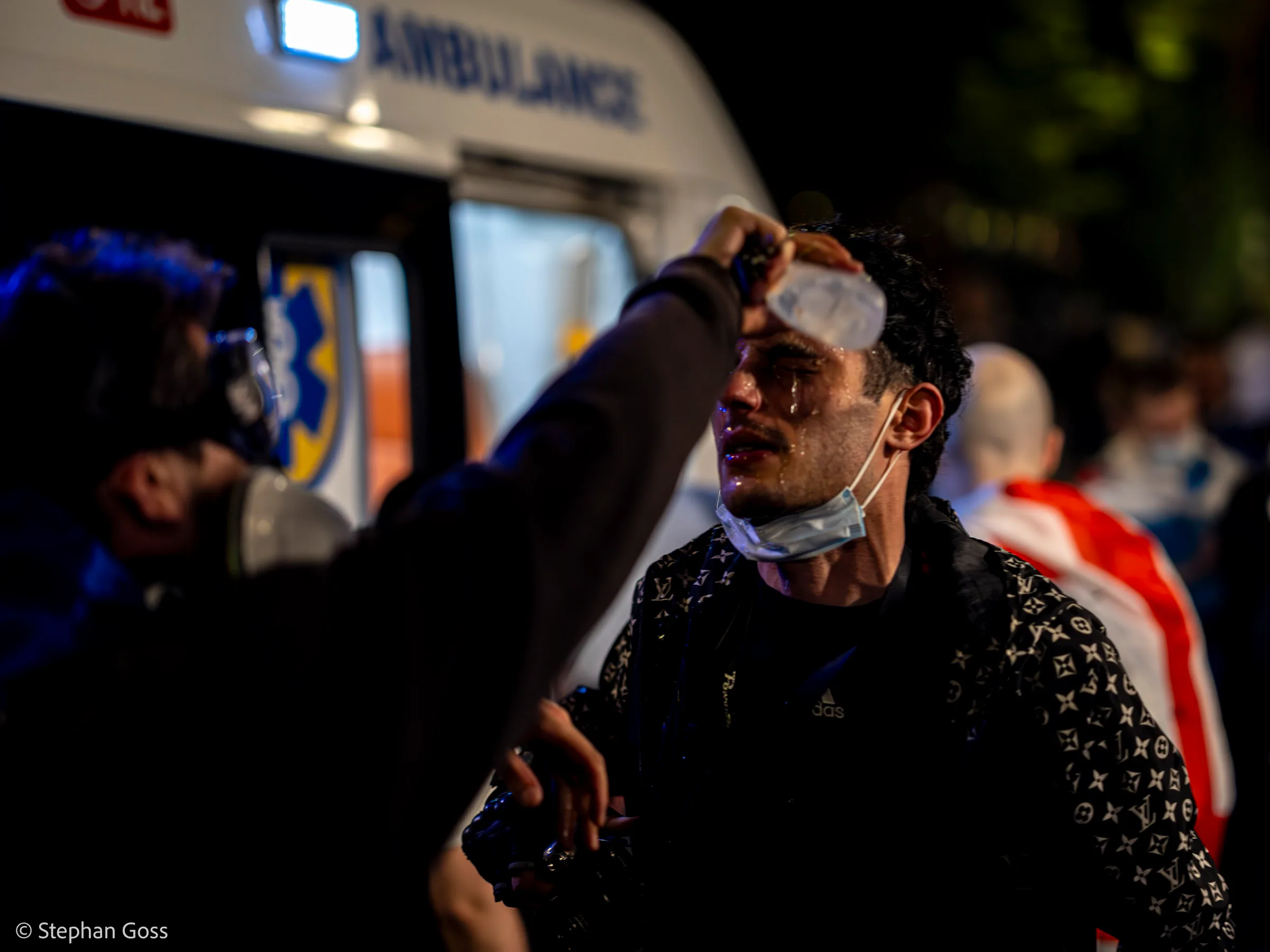 A young man with a dressing taped to his forehead and tears running down his face stands in front of an ambulance while a medic reaches up to hold the bandage in place.