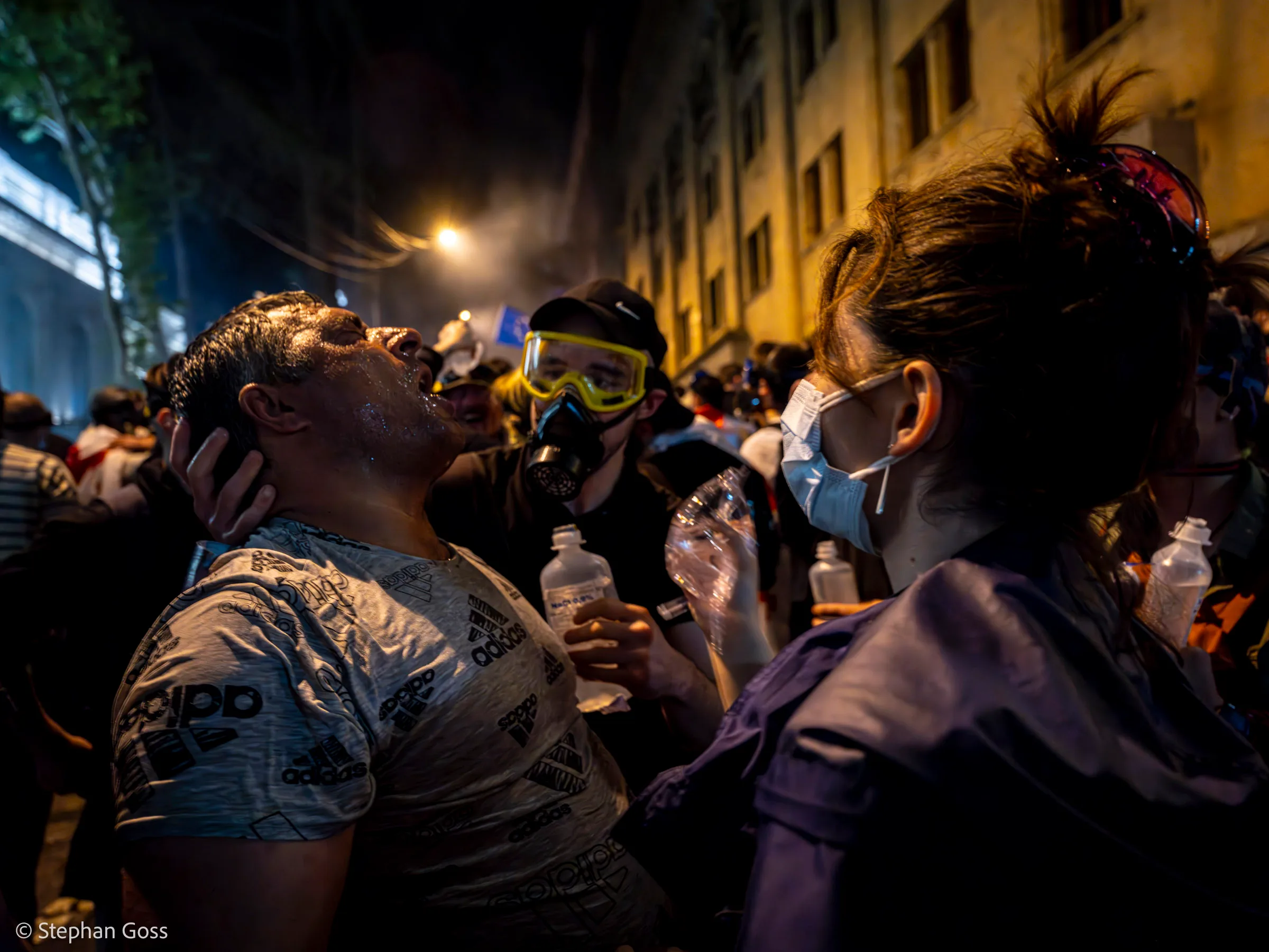 A man tilts his head back with his eyes shut while a woman in a face mask steadies him and pours water into his eyes; others stand ready with more bottles.