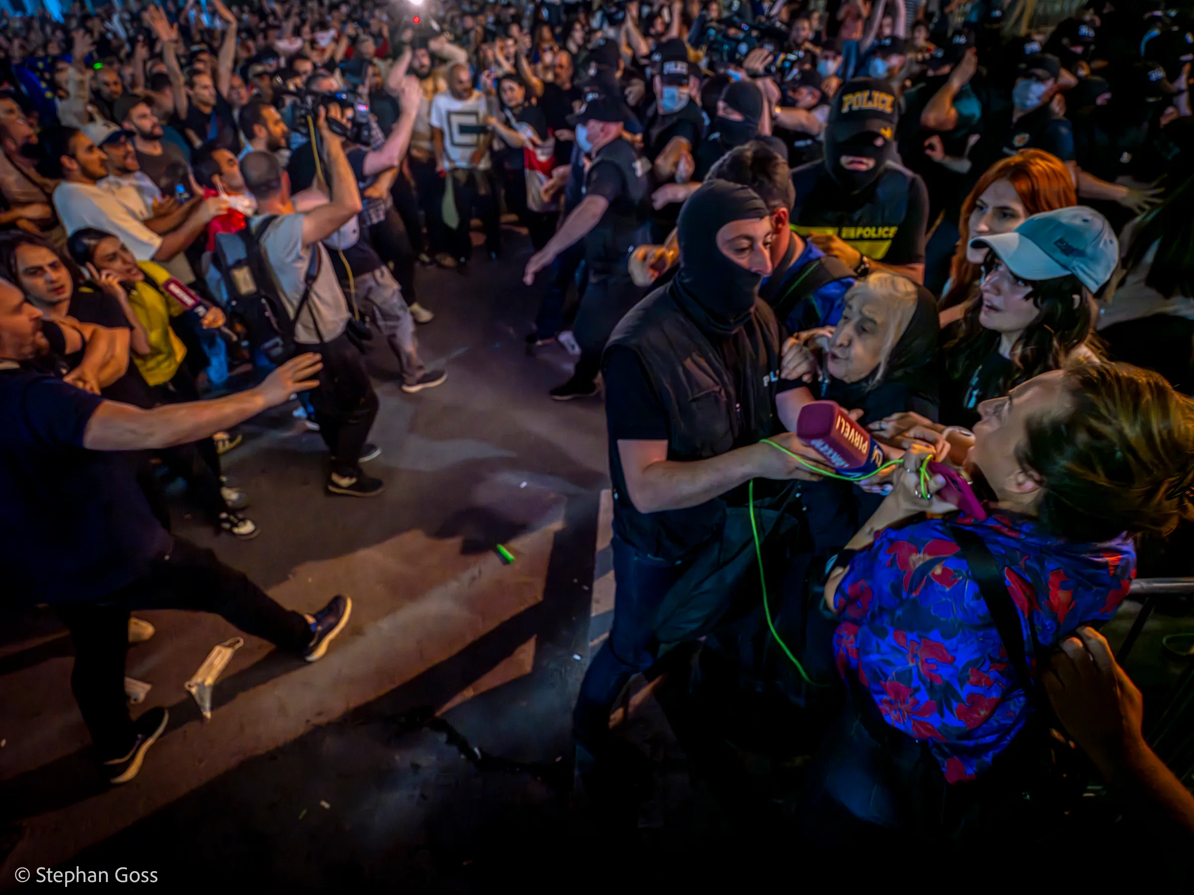 Masked officers in black seize demonstrators at the front of a crowd; an elderly woman is caught between them as the people around her shout and reach forward.