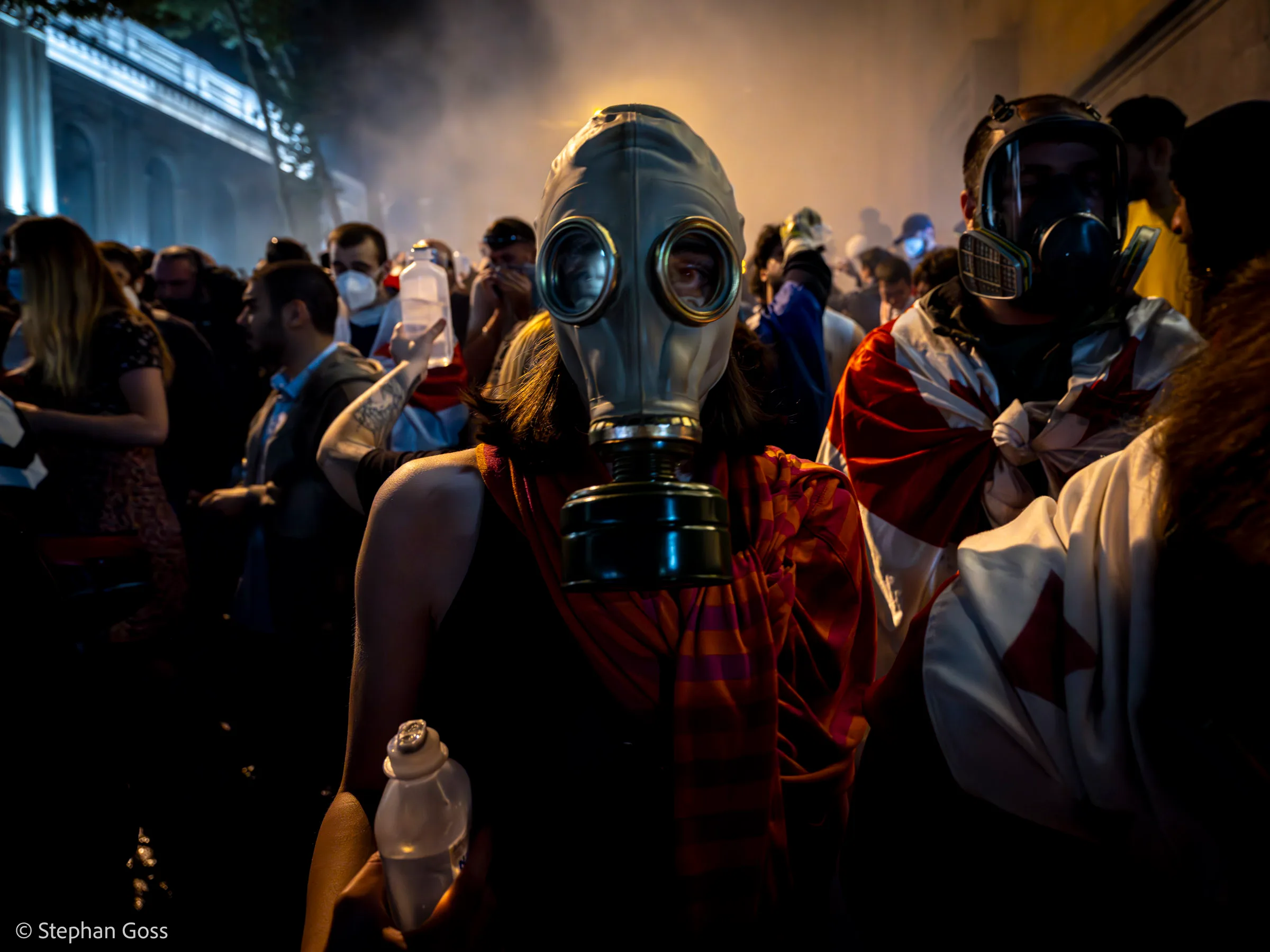 A protester in a Soviet-pattern gas mask stands in drifting tear gas with a Georgian flag over their shoulders, holding a water bottle; another masked demonstrator wrapped in a flag stands behind.