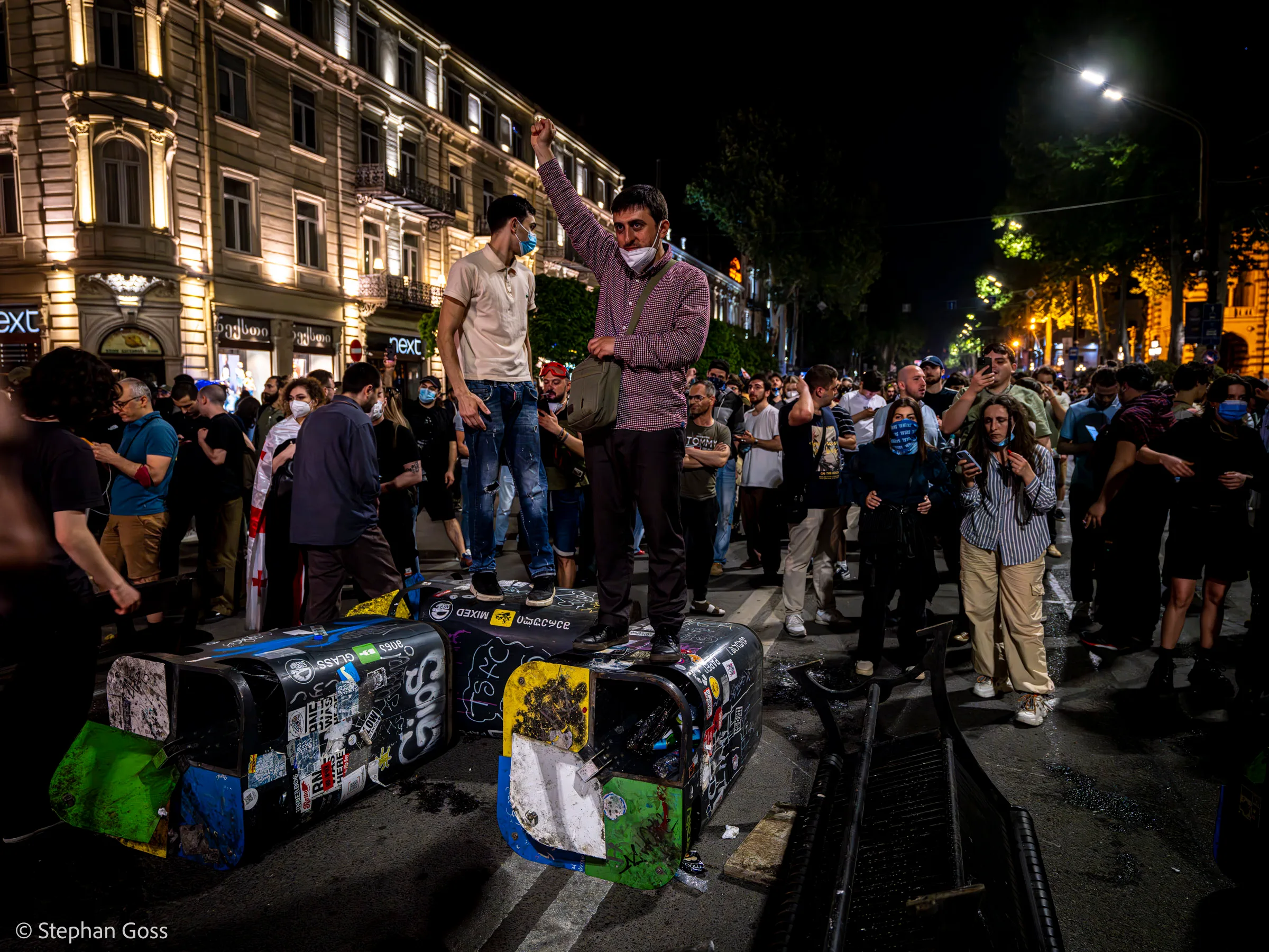 Two men stand on a row of sticker-covered wheelie bins dragged into the road; one raises a clenched fist above a crowd that fills the street behind him.