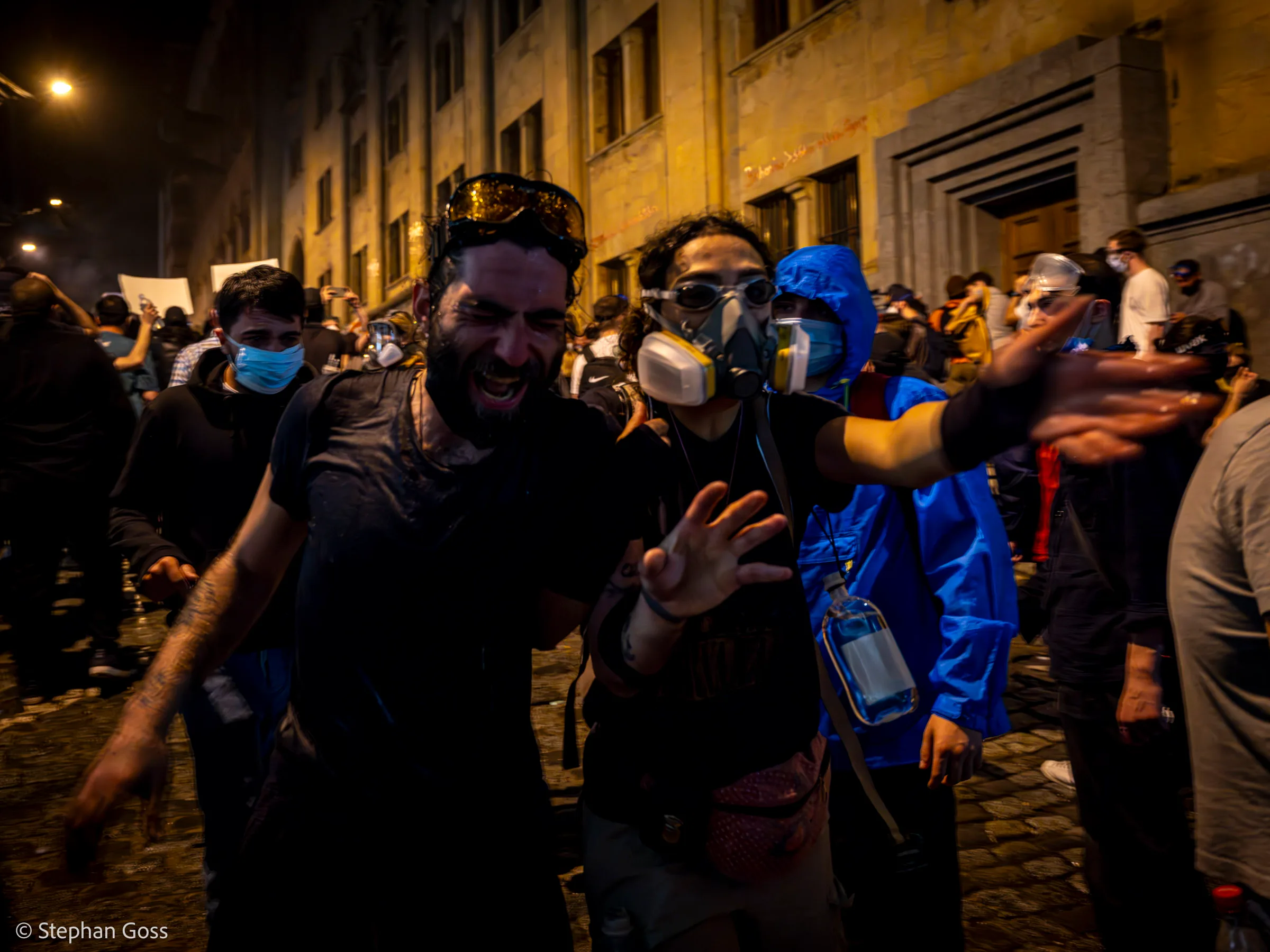 A man in a black T-shirt shouts with his arm outstretched, a woman in a gas mask beside him and a volunteer in a blue protective suit behind.