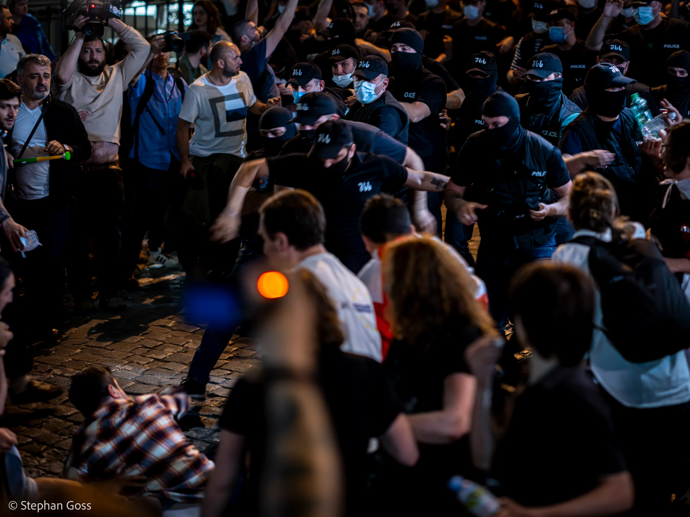 A line of masked riot police in black push into a crowd on a cobbled street at night; a man in a white T-shirt is shoved backwards while onlookers film on their phones.