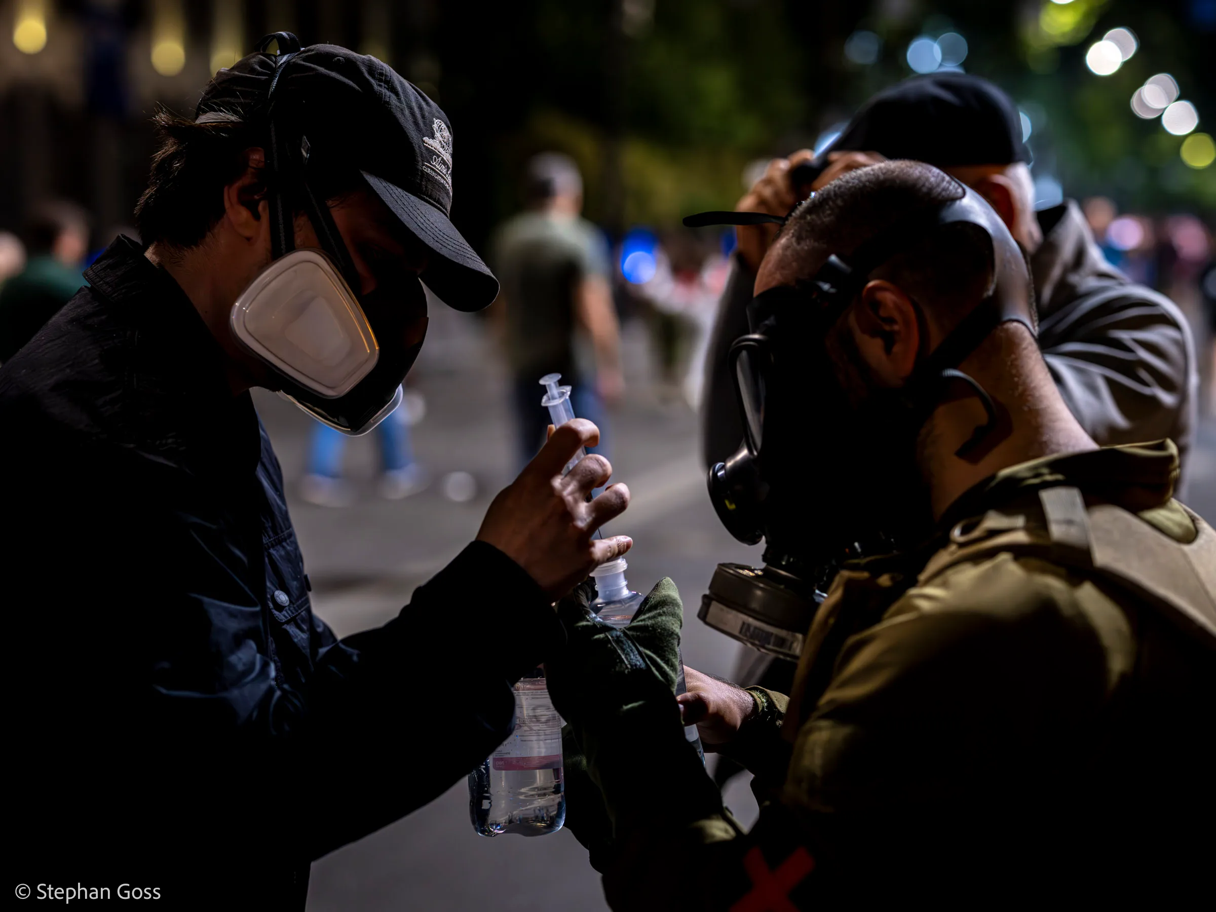 A man in a cap and white respirator hands a water bottle to a volunteer medic in a full gas mask wearing a red cross armband, on a street lit by street lamps.