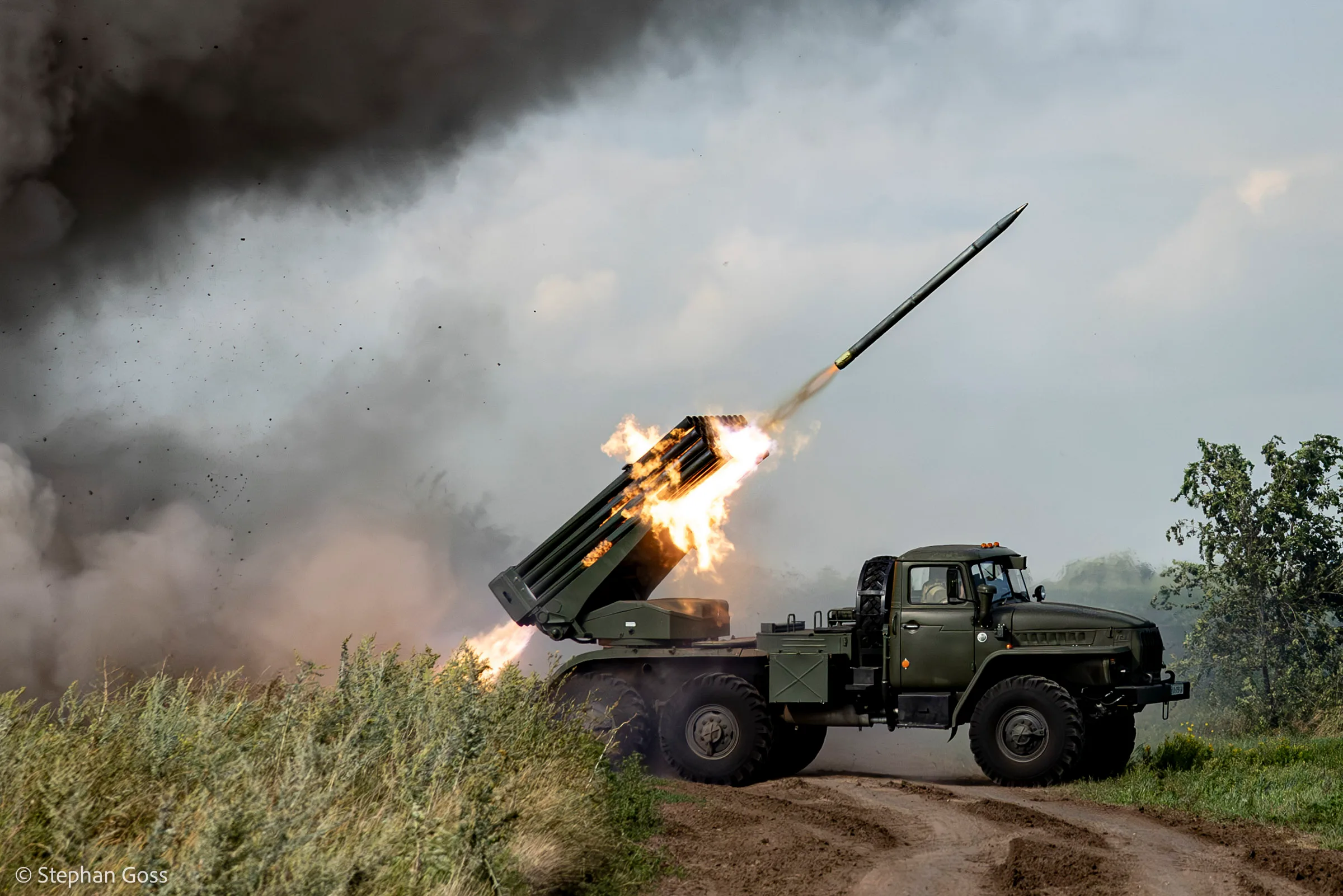 A BM-21 Grad multiple rocket launcher fires from a dirt track at the edge of a field, flame and black smoke pouring from the tubes as a rocket clears the barrels.