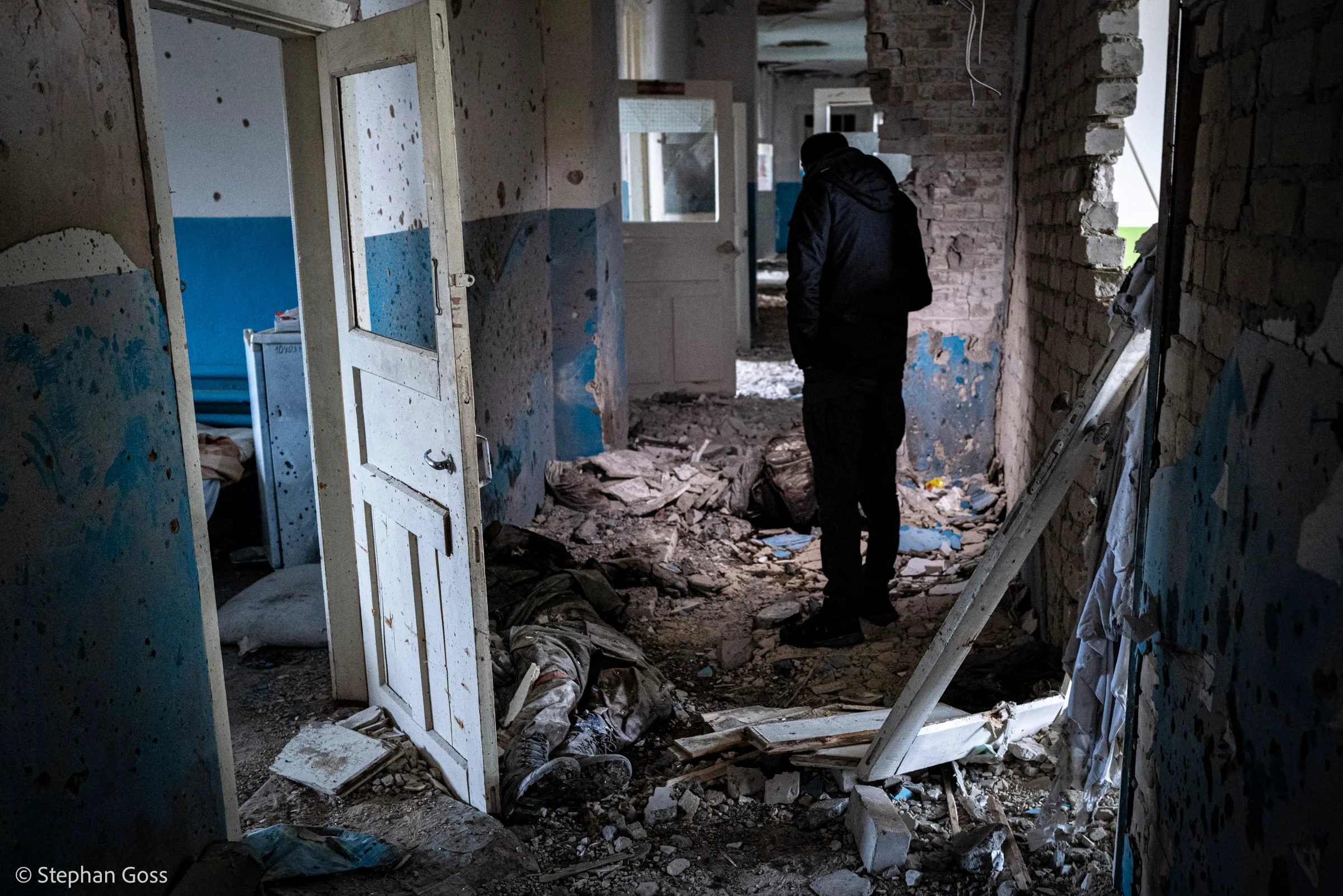 A man stands in the wrecked interior of a public building, the corridor ahead of him filled with rubble, the walls pocked by shrapnel and the doors blown off their frames.