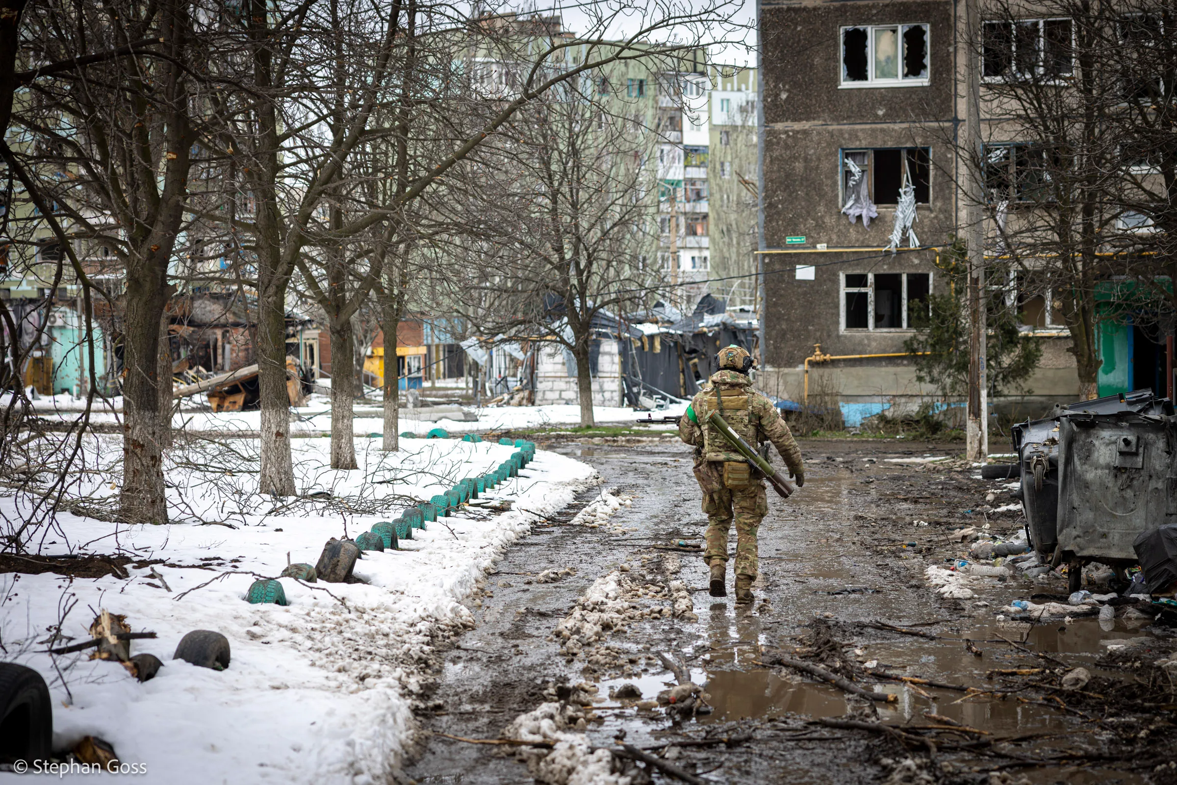 A Ukrainian soldier walks alone down a slush-covered road between burnt-out apartment blocks, his rifle at his side, under bare winter trees.