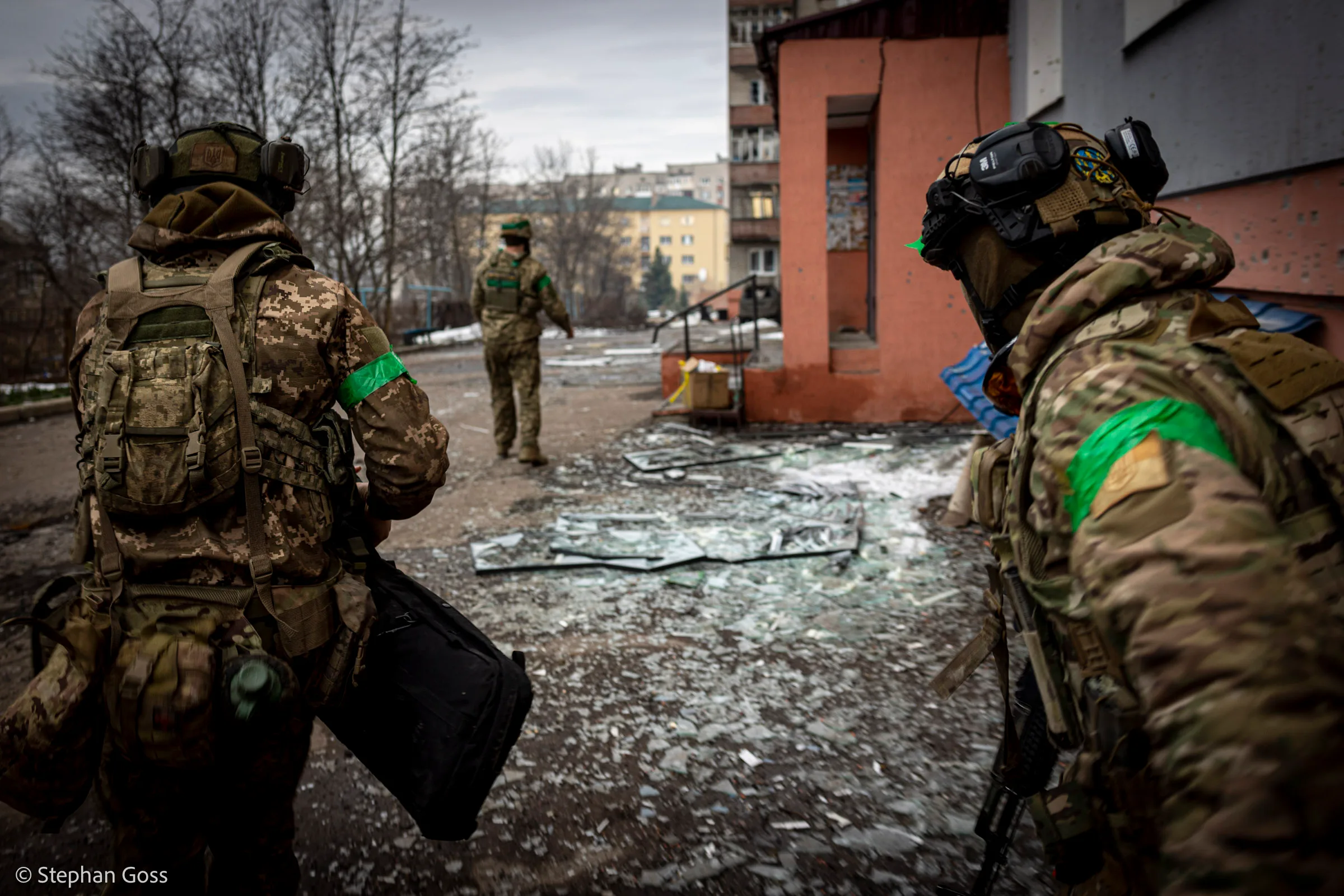 Two soldiers in camouflage with green identification armbands move towards a damaged apartment block across ground carpeted with shattered glass.