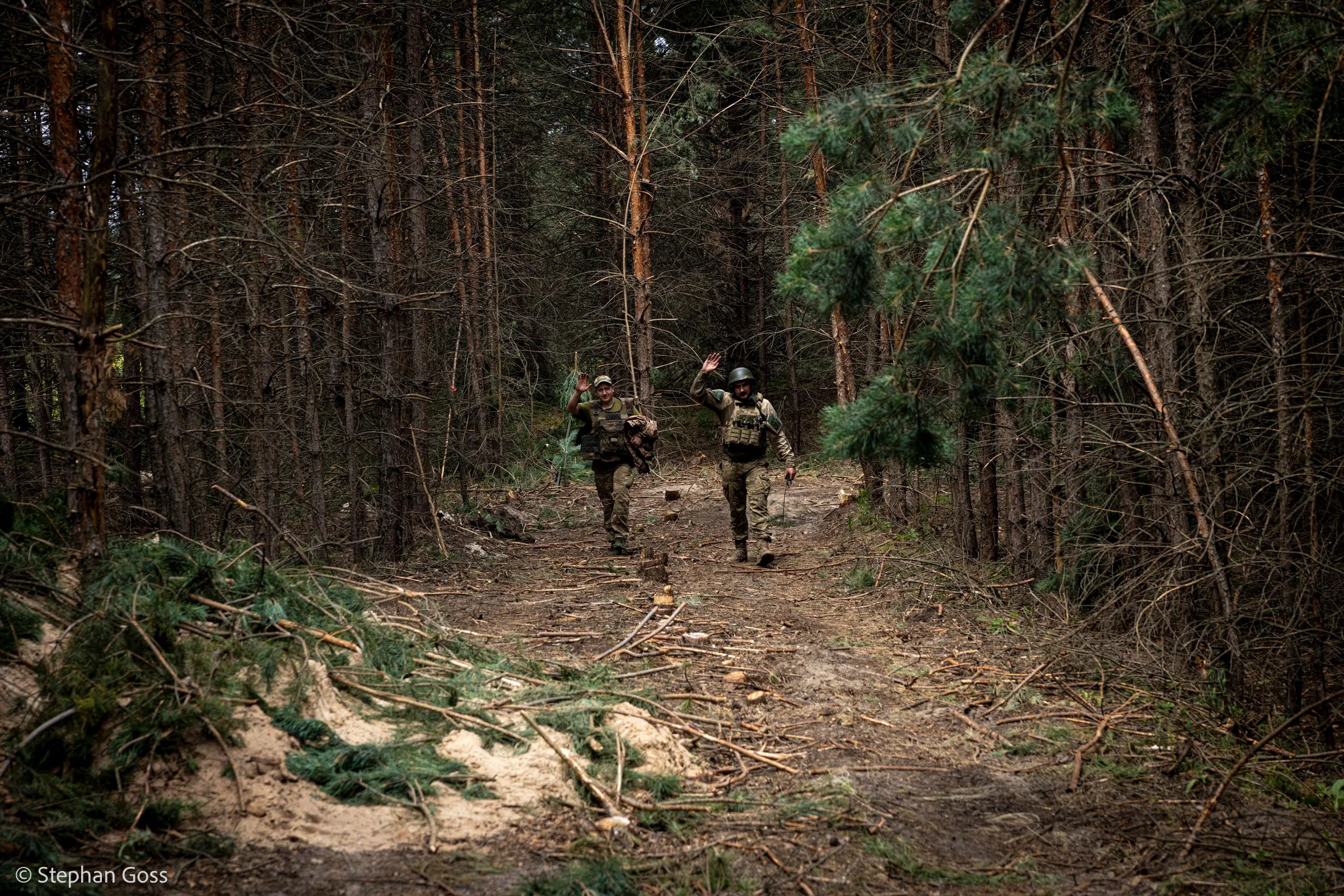 Two soldiers walk up a sandy track through dense pine forest, cut branches and camouflage netting piled along the roadside.